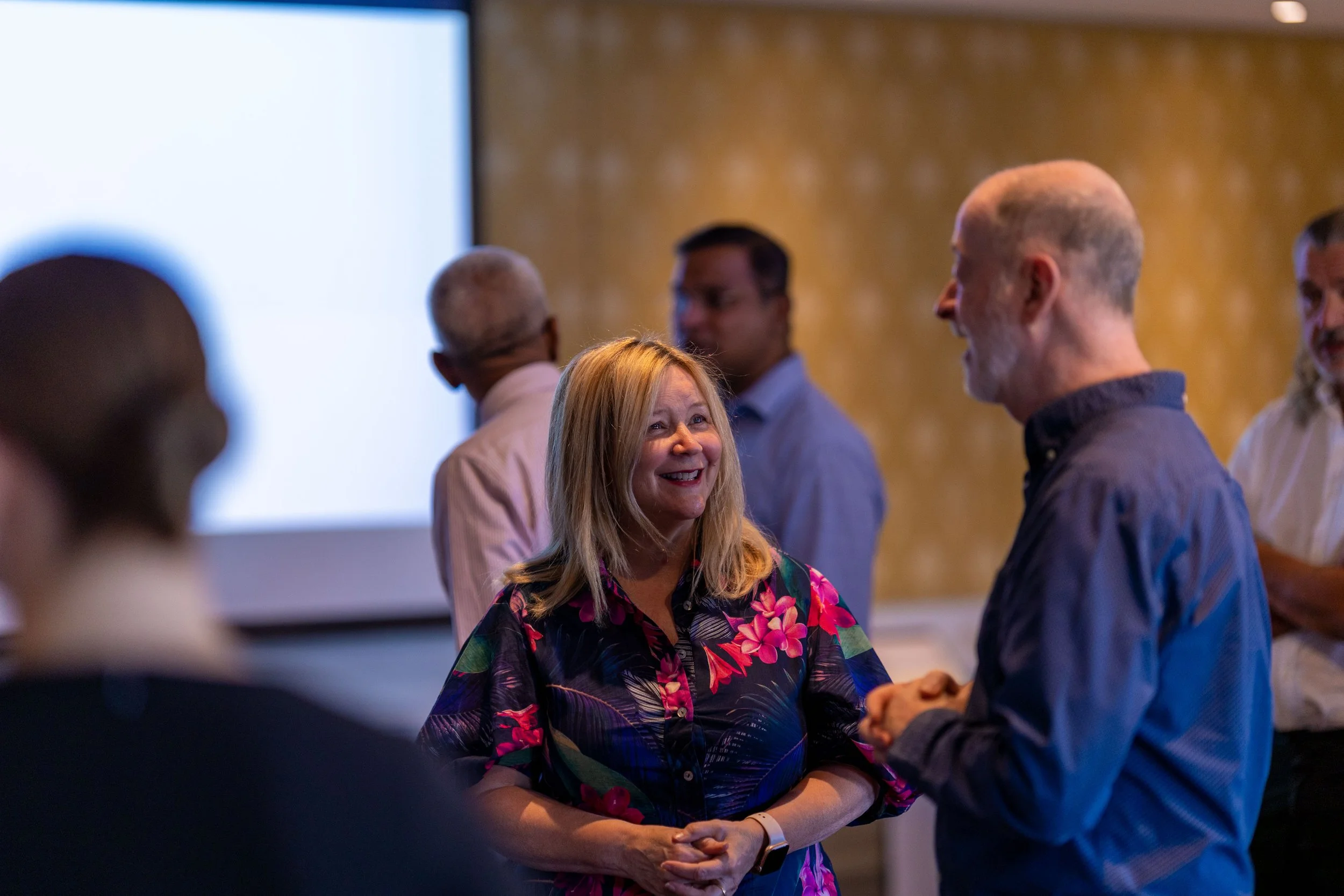 A group of people at a social gathering, with a woman with blonde hair wearing a colorful floral shirt smiling and talking to a man with short gray hair in a dark shirt, while others are engaged in conversation in the background.