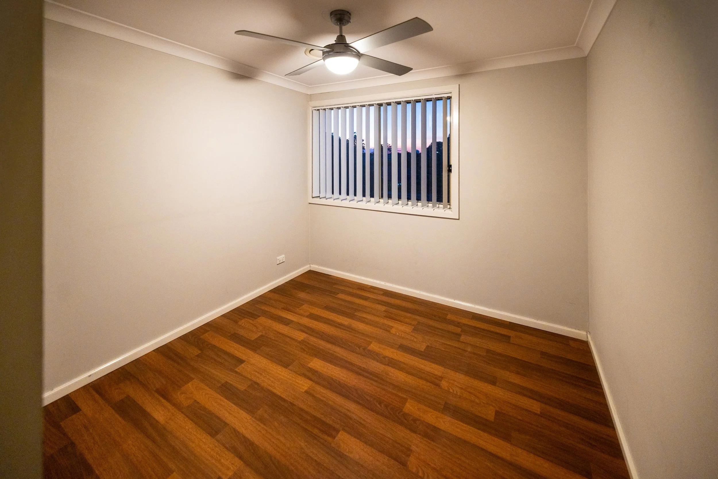 Empty room with white walls, wooden floor, a ceiling fan with light, and a window with vertical blinds showing an evening sky.
