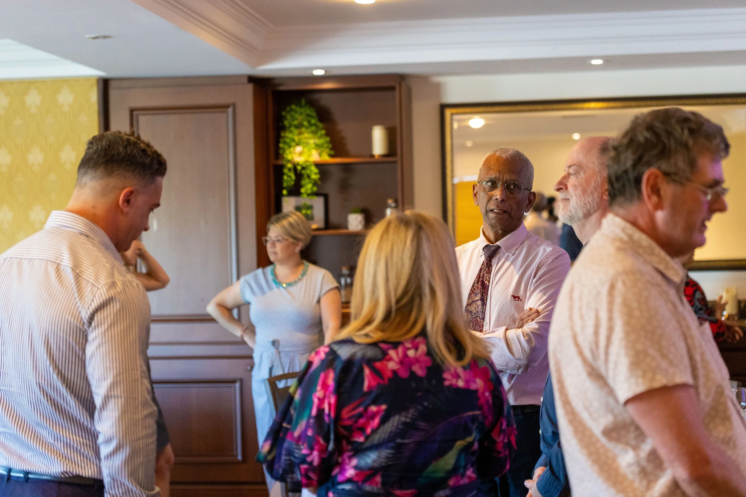 Group of people engaged in conversation in a room, some standing near a dark wood cabinet with decorative items and a mirror.