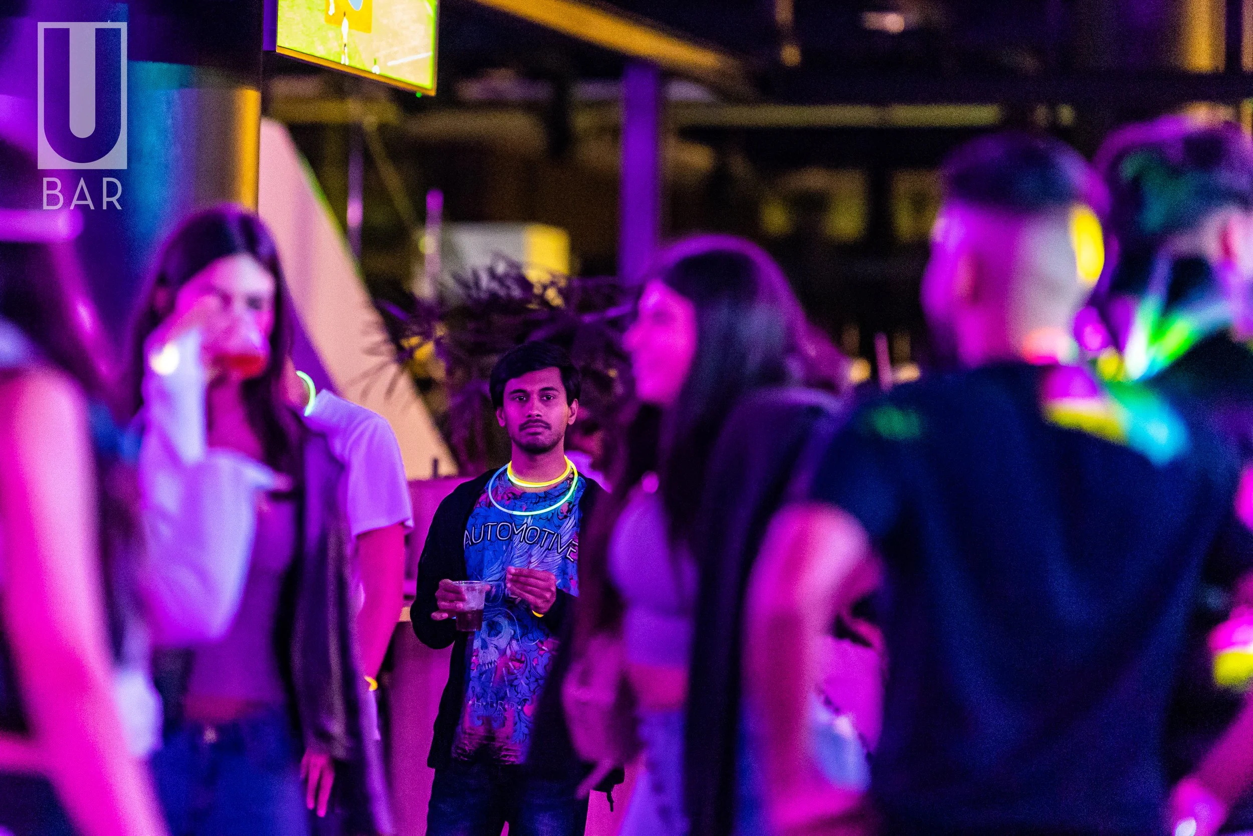 A group of young adults socializing at a bar with neon lighting, with one man standing in the center holding a drink and looking directly at the camera.