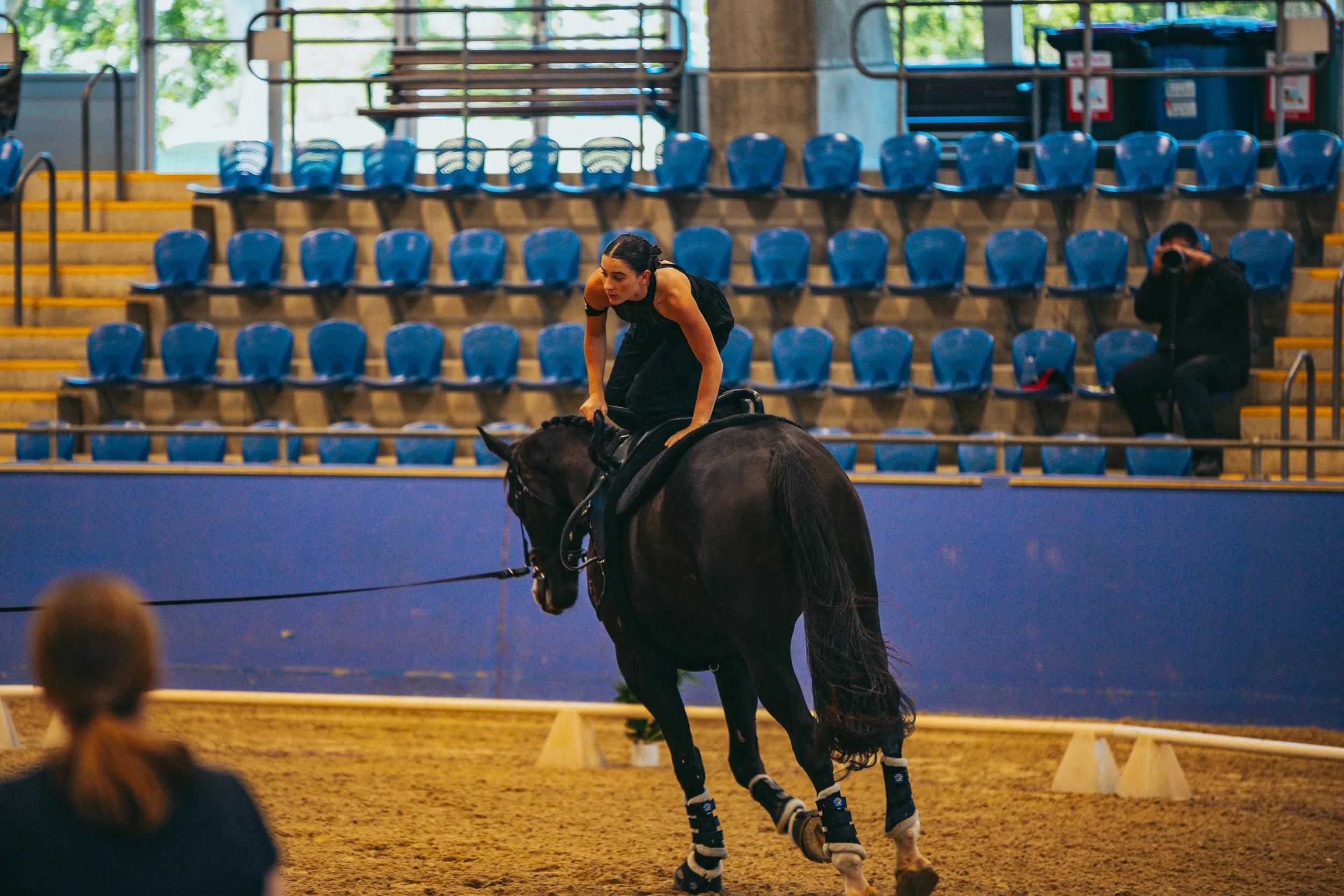 A woman dressed in black riding a dark horse in an indoor arena with blue seats in the background and a person sitting on the stairs taking photos.