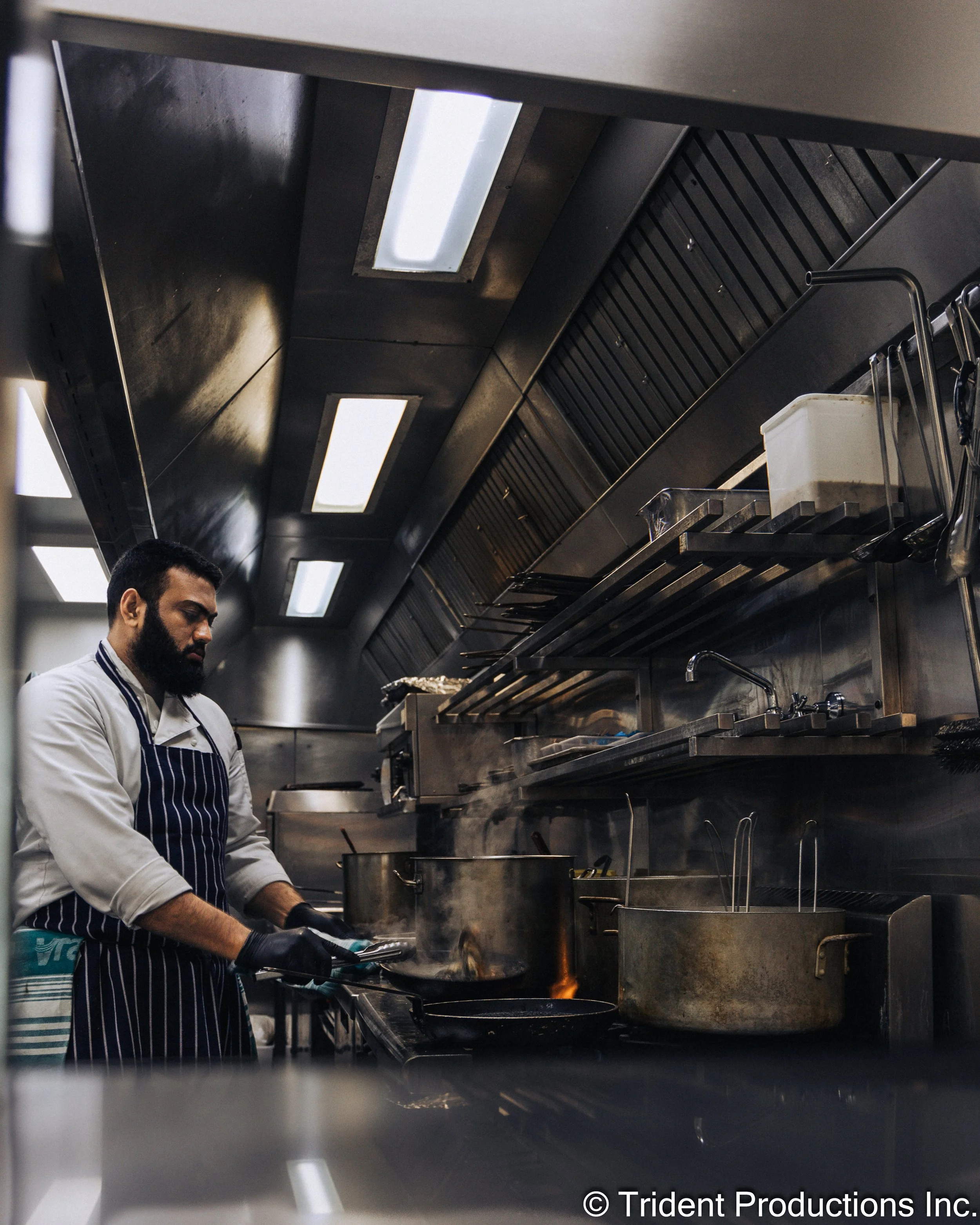 A chef with a beard, wearing a white shirt and blue-striped apron, cooking on a stove in a professional kitchen. The kitchen has stainless steel surfaces and equipment, with pots and utensils visible.