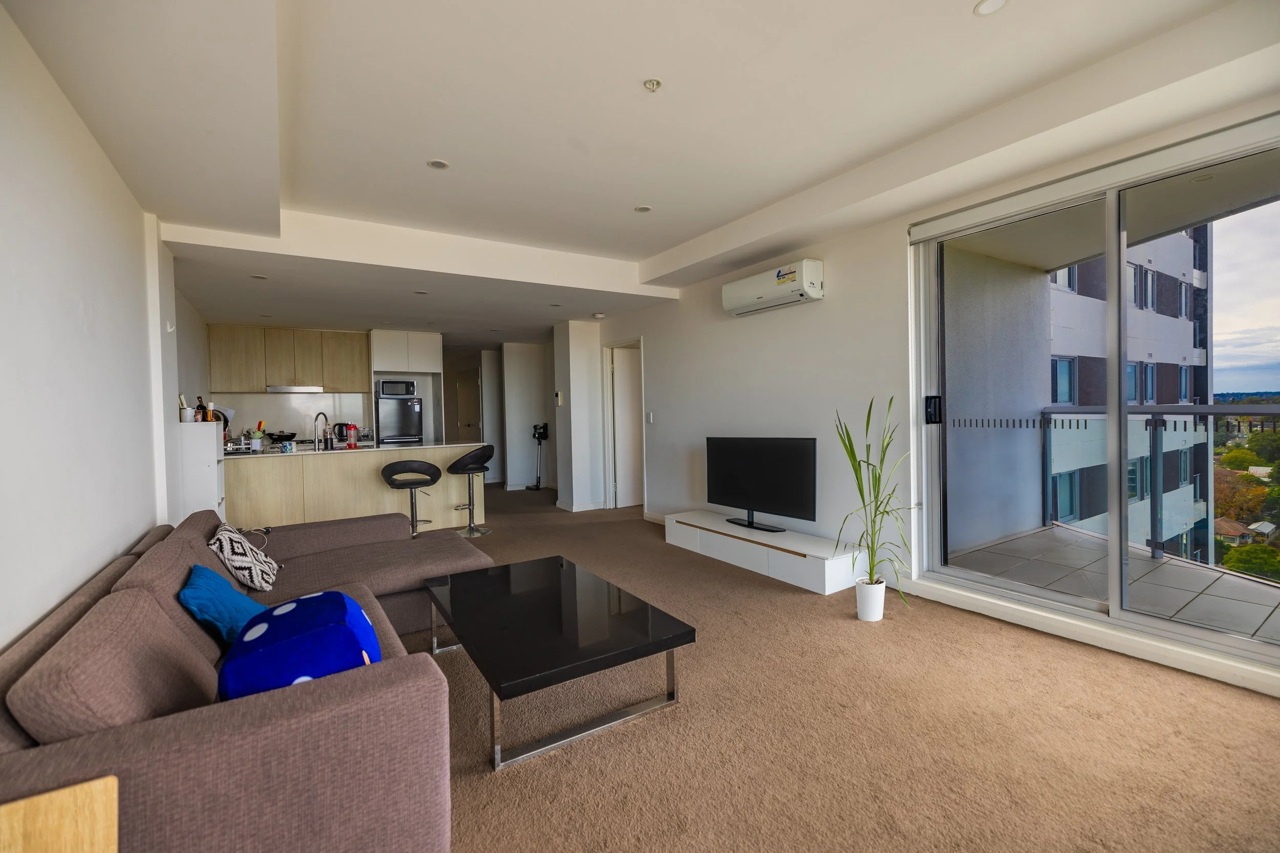 Living room with brown sofa, black coffee table, TV on white stand, potted plant, sliding glass door leading to balcony, open kitchen in background with bar stools, and city view outside.
