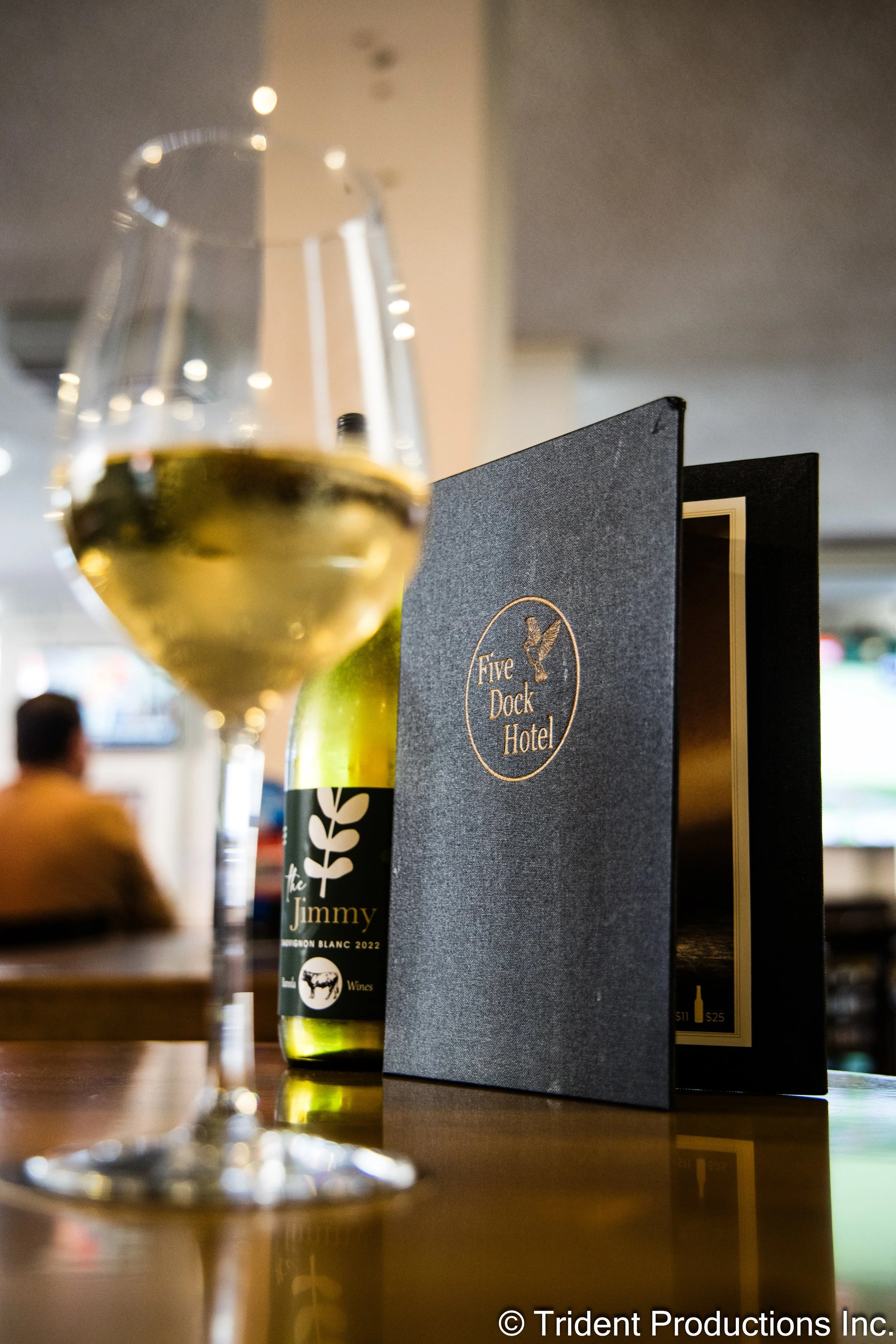A wine glass with white wine on a restaurant table, with a bottle of wine and a menu from Five Dock Hotel in the background.
