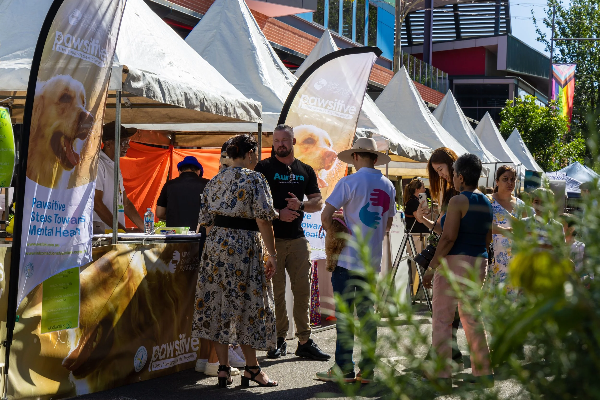 People at an outdoor event with tents, likely a health fair or community gathering. Several people are talking and taking photos near booths with banners and informational signs.