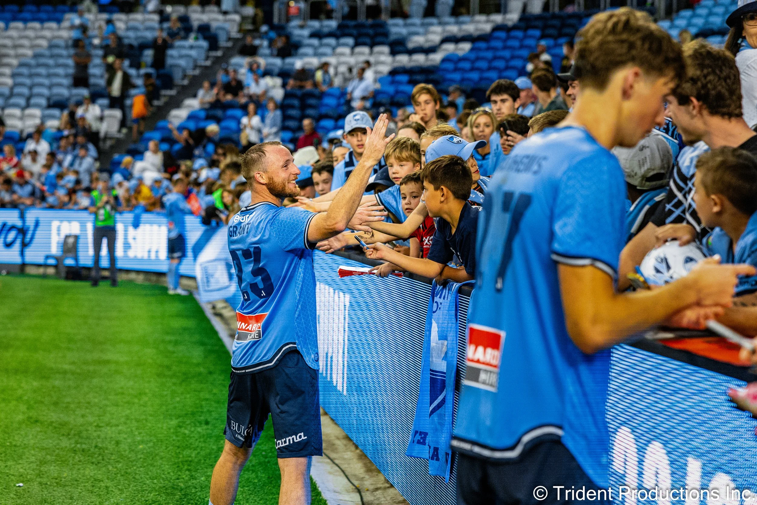 Soccer player in blue jersey interacting with young fans behind the sideline fence at a stadium with empty blue seats in the background.