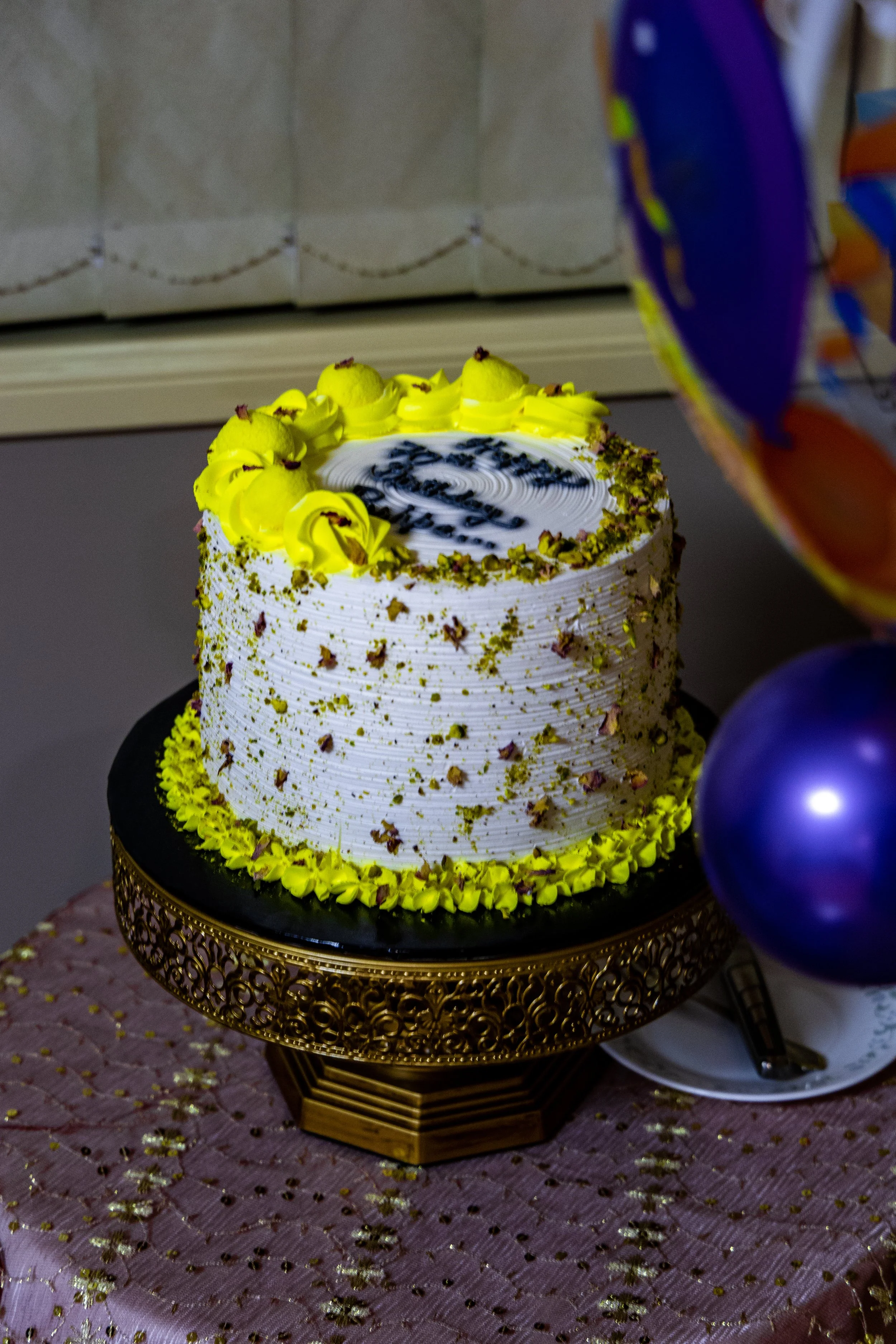 A two-tier white birthday cake decorated with yellow and green accents, placed on a decorative gold stand with a pink tablecloth.