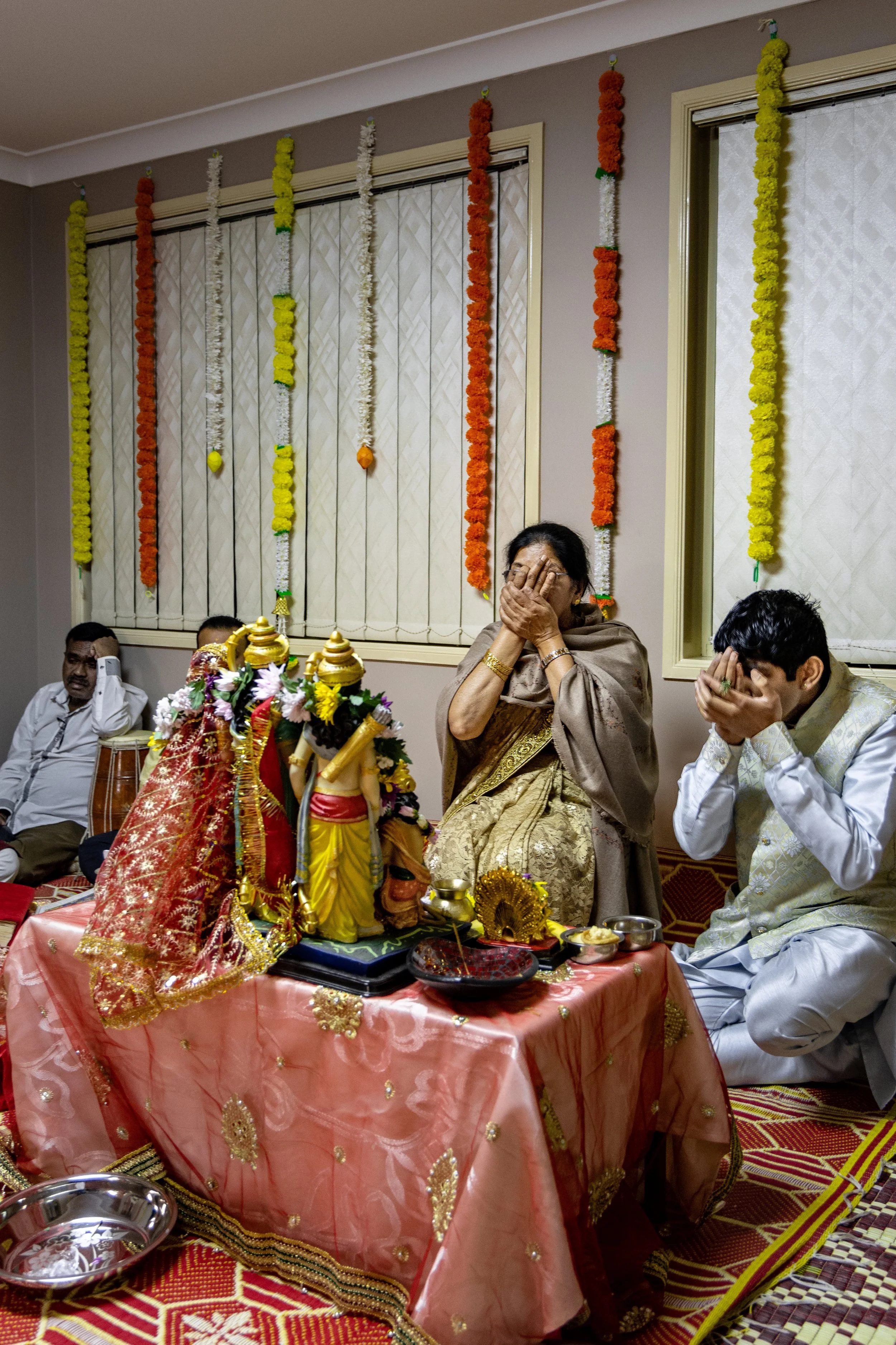 People participating in a Hindu religious ceremony, sitting at a decorated altar with statues and offerings, inside a room decorated with marigold garlands.