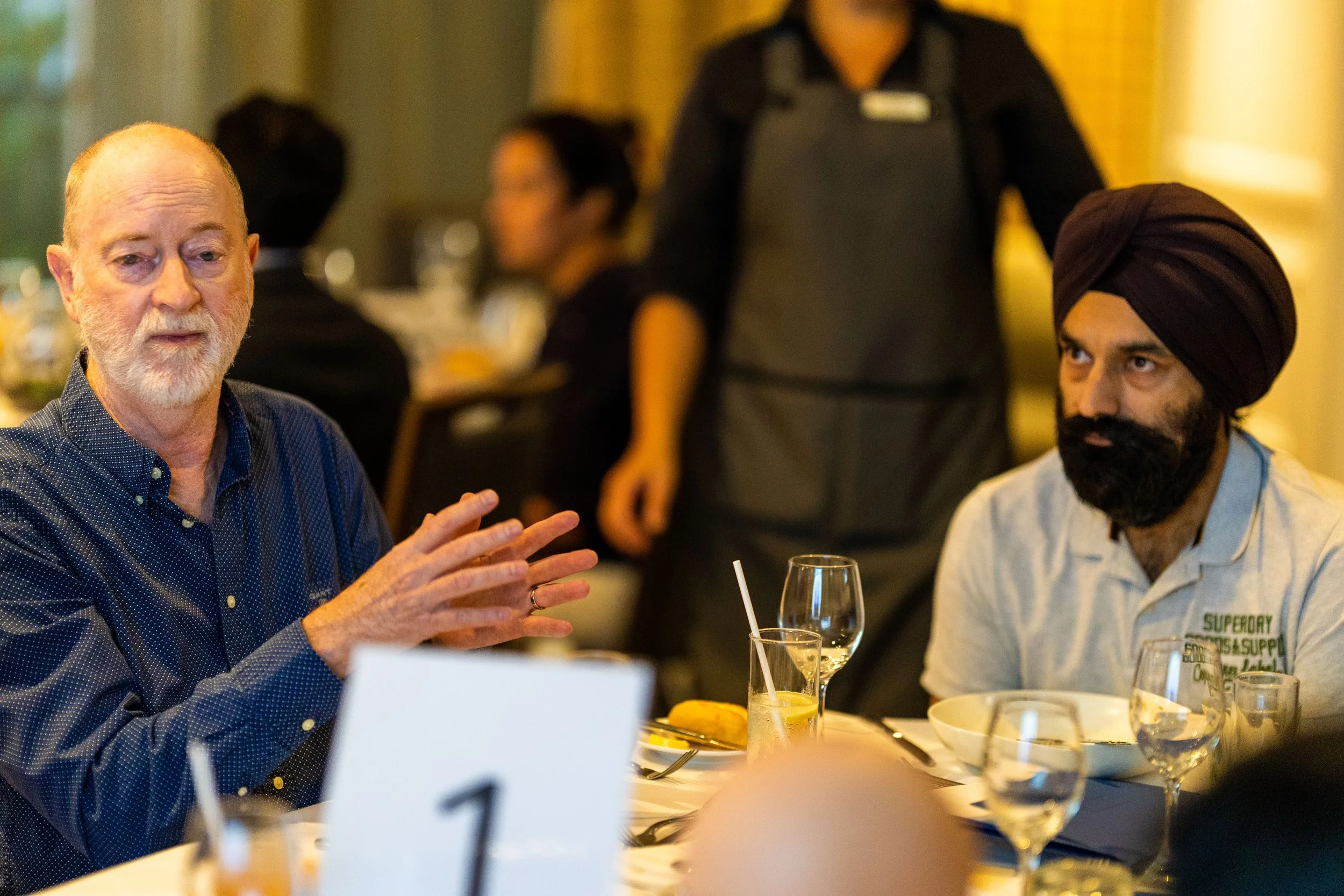Two men sitting at a dining table having a discussion, with a waiter in the background, in a restaurant setting, with glasses and plates on the table.