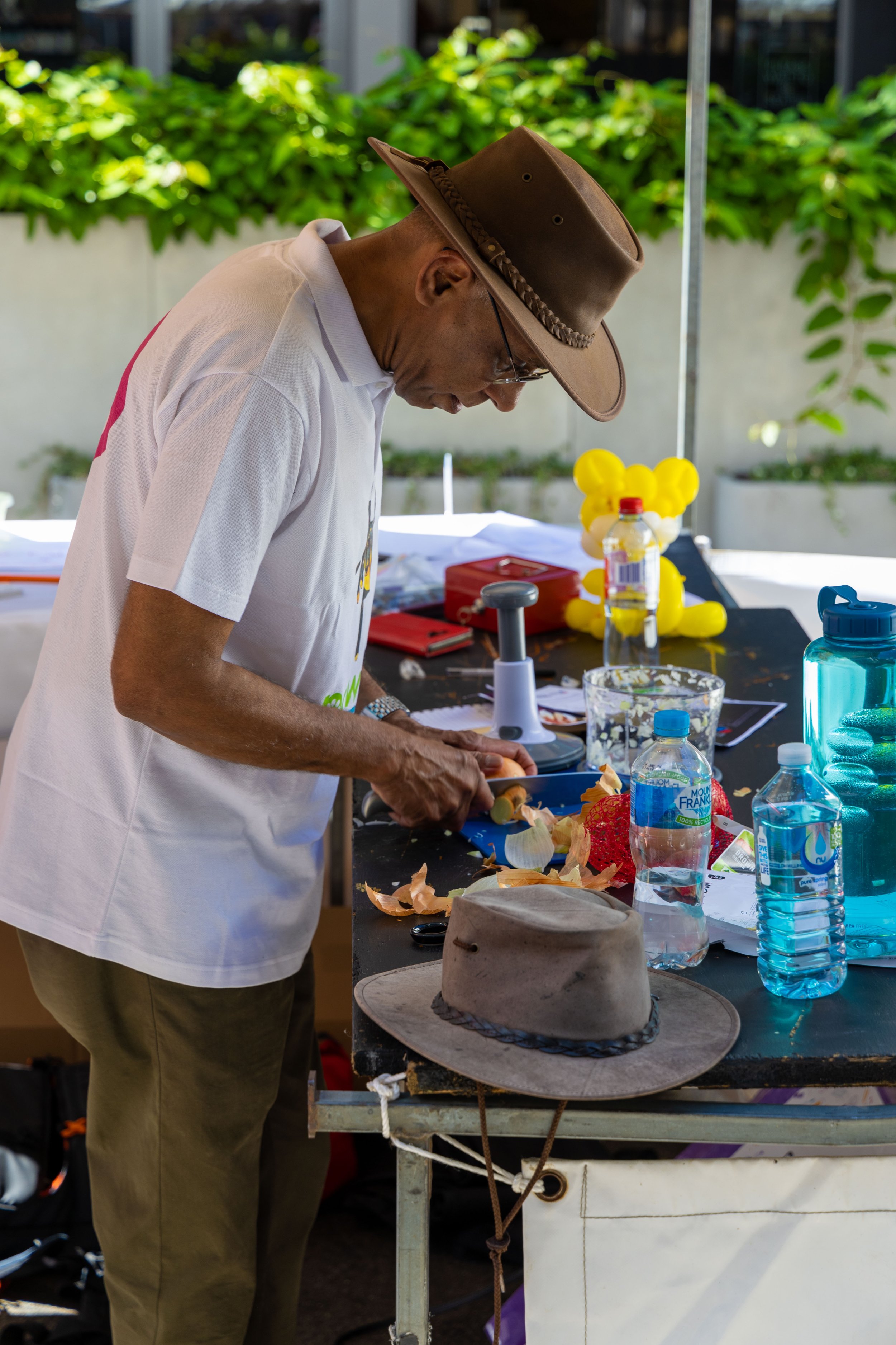 A man wearing a cowboy hat, glasses, and a white t-shirt is chopping vegetables at a table outdoors, with a background of green foliage.