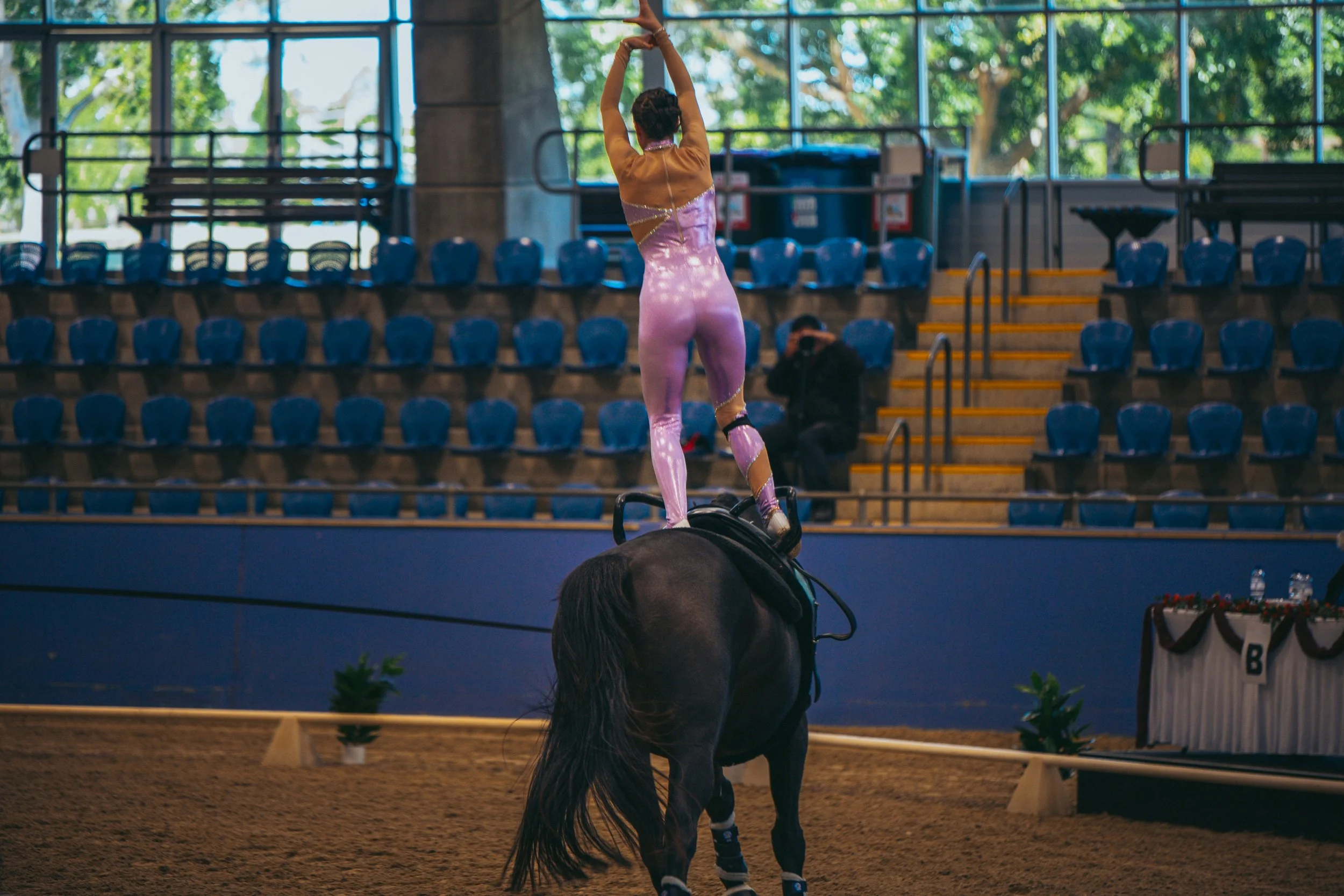 A gymnast in a shiny pink and purple leotard practices balancing on a horse in an indoor arena with blue seats and large windows.