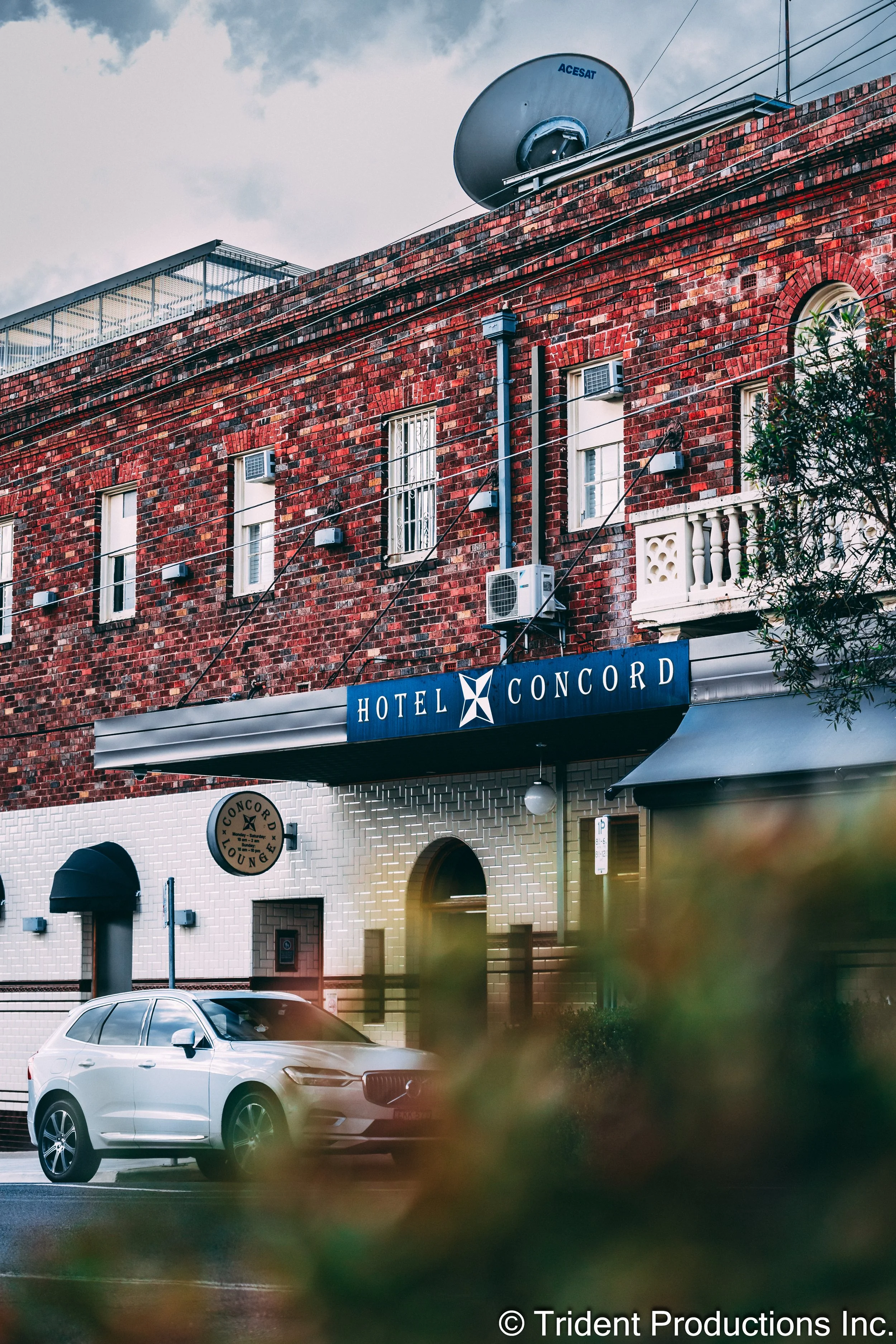 Exterior view of the Hotel Concord, a brick building with a blue hotel sign, windows, air conditioning units, satellite dish, parked white car, and greenery in the foreground.