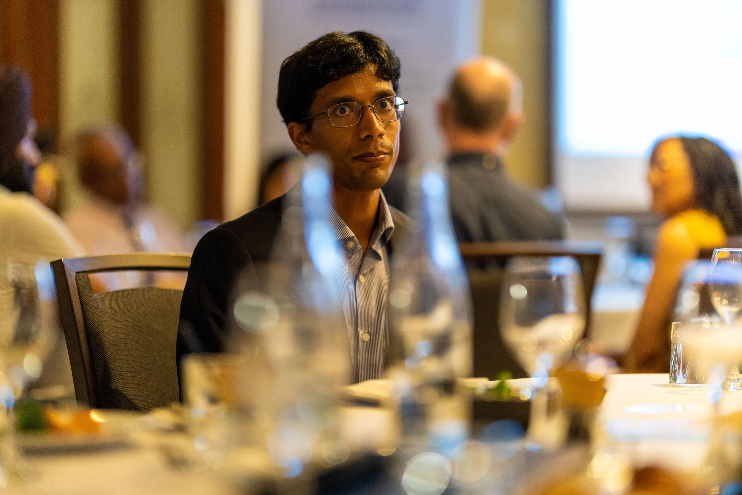 A man with dark hair, glasses, and a suit sitting at a banquet table in a dimly lit room, with other people seated around.