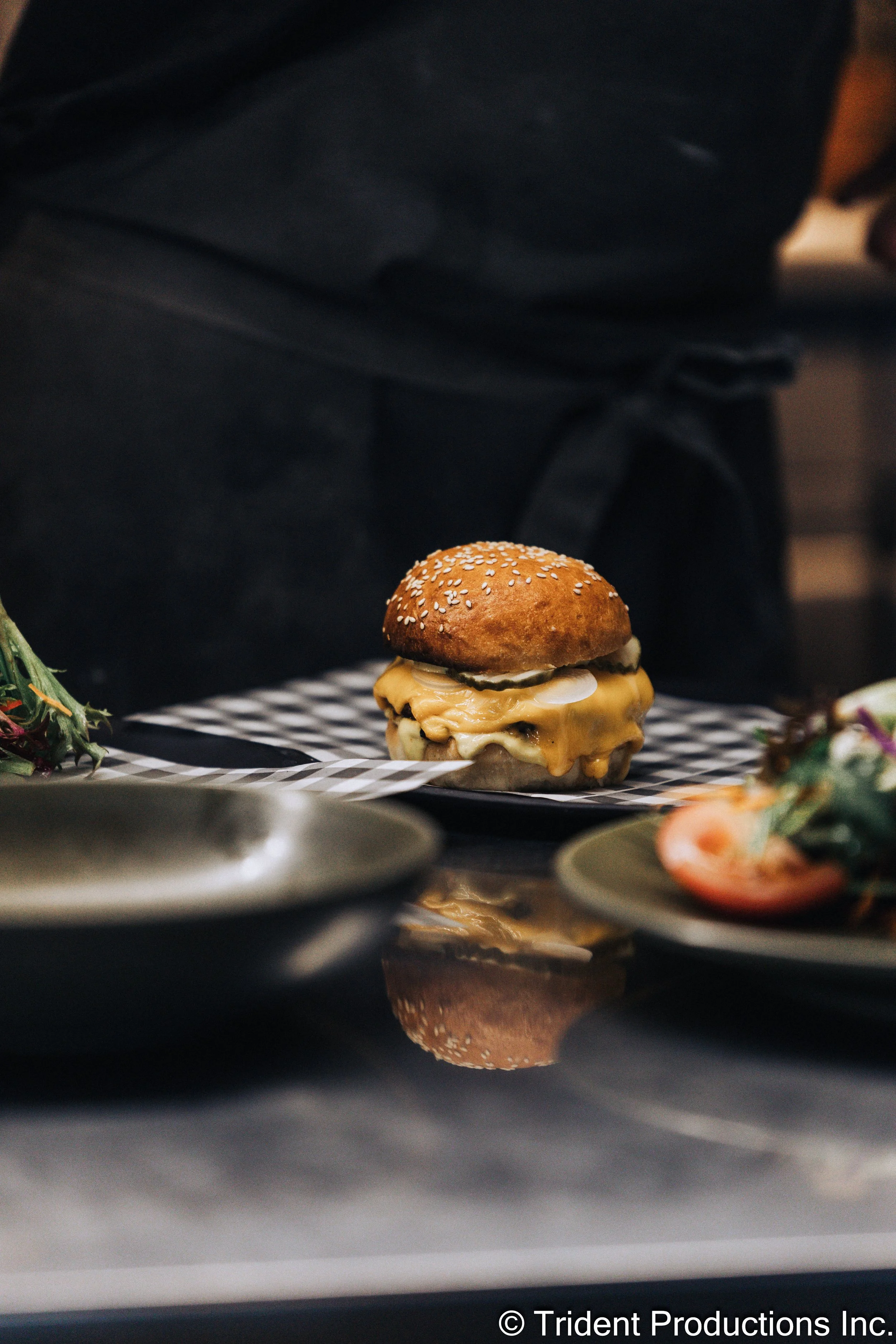 A cheeseburger with melted cheese, pickles, and a sesame seed bun on a black and white checkered paper, placed on a tray.