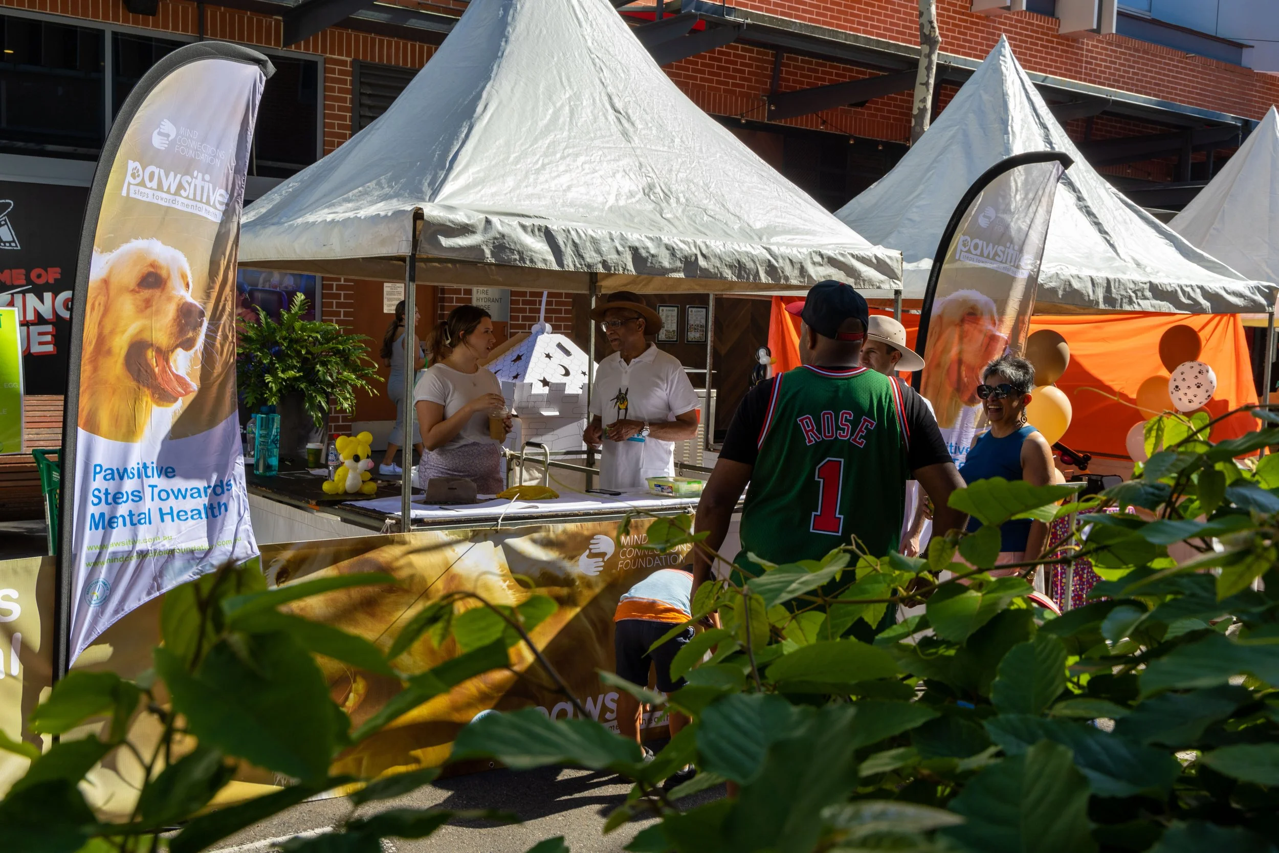 People at an outdoor event with tents promoting mental health awareness, including a booth with a banner showing a golden retriever dog and the text 'Pawsitive Steps Towards Mental Health'.