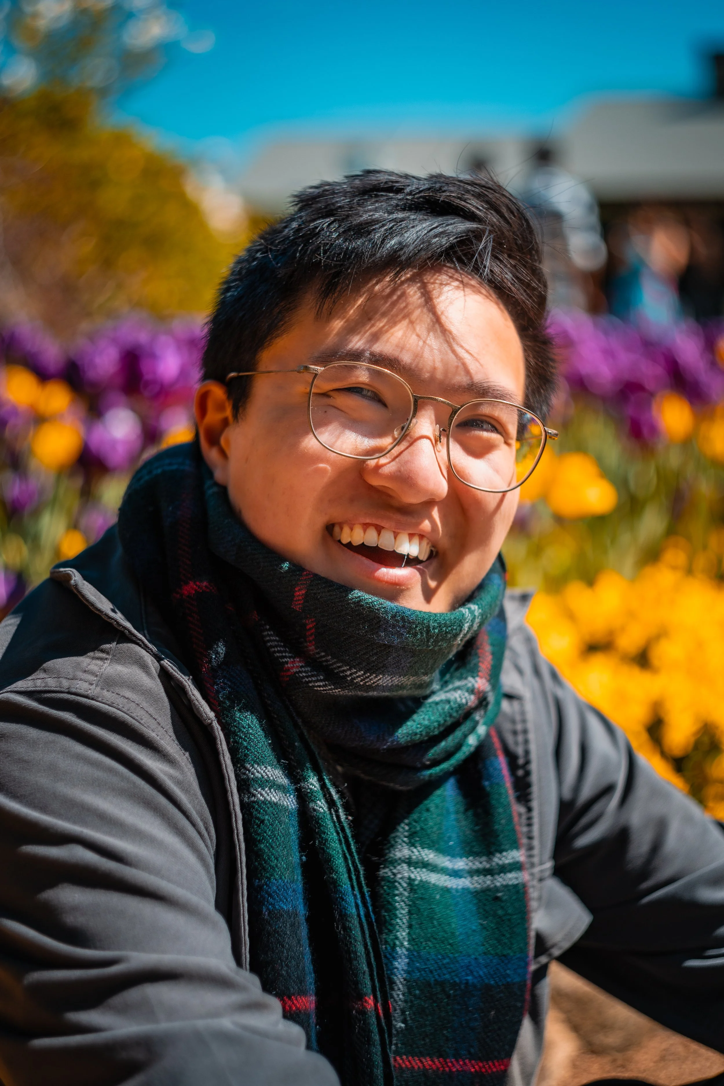 Smiling person with glasses and a plaid scarf outdoors with colorful flowers in the background.