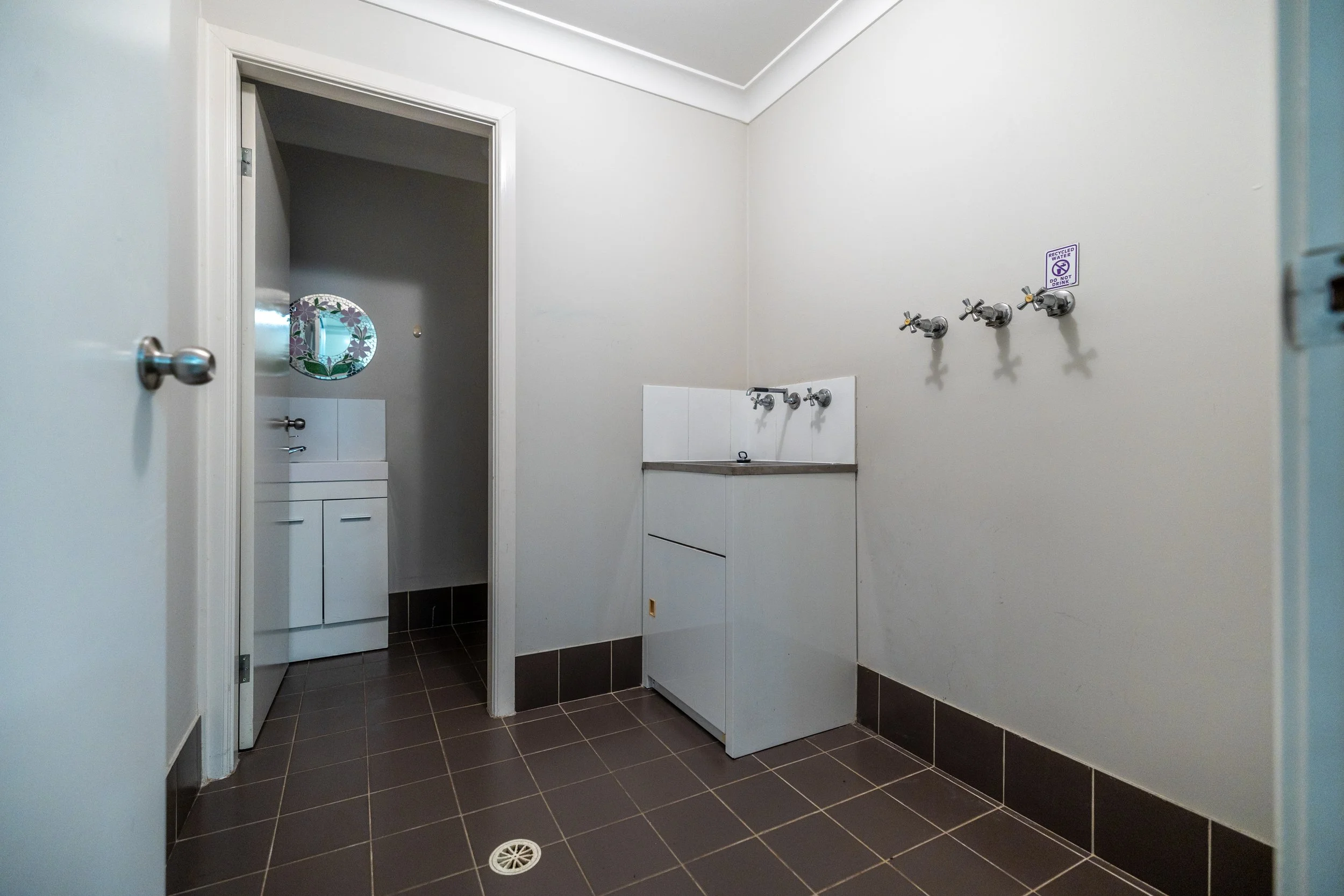 Empty laundry room with brown tiled floor, white walls, a small white sink unit, and three plumbing hookups on the wall.