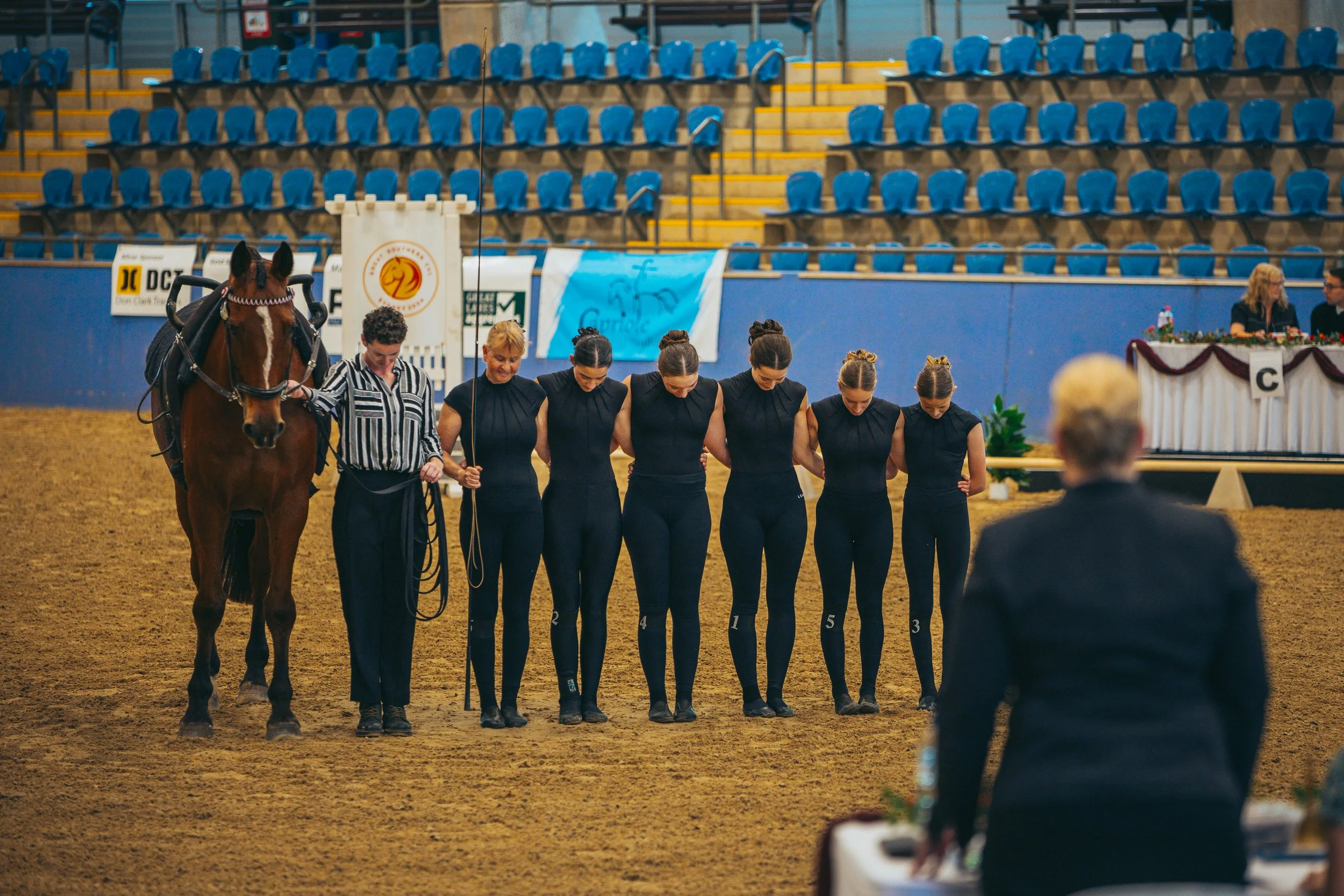 Six women in athletic attire bowing with a horse and trainer in an indoor arena during an equestrian event.