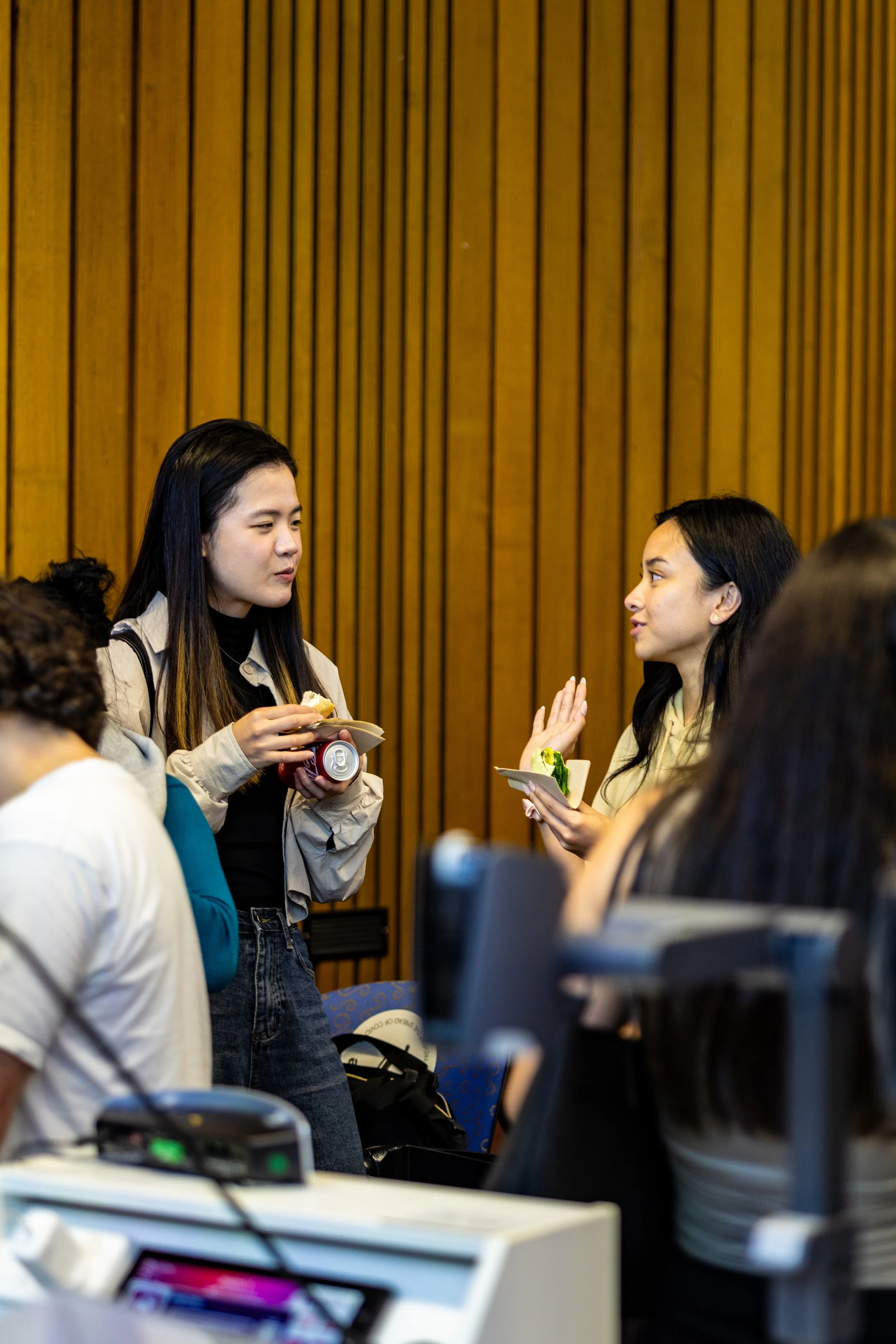 Two women standing and talking at an indoor event or conference, one holding a plate and drink, the other gesturing with food on her plate, with wooden paneling background.