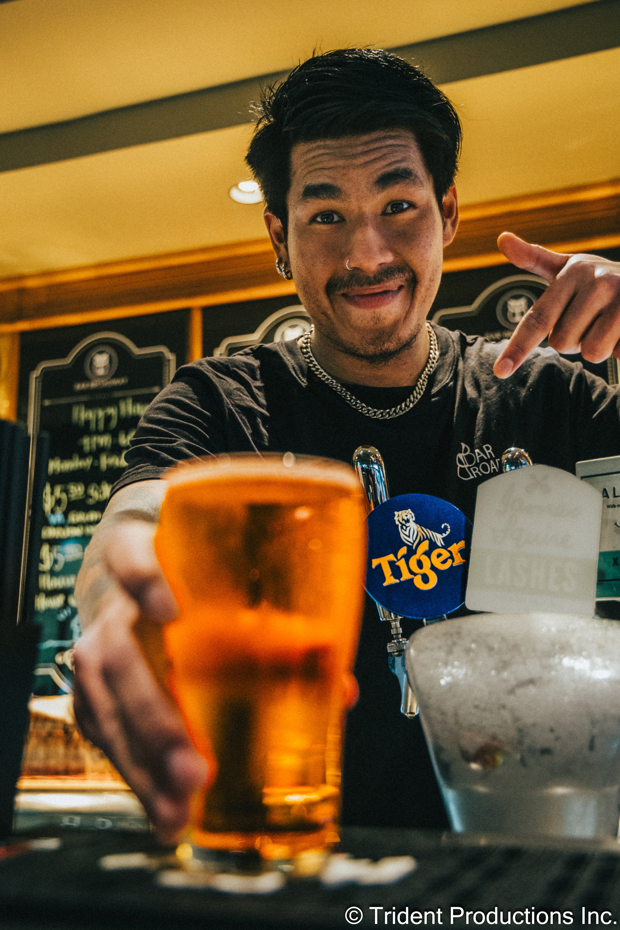 A man with black hair, a mustache, and facial hair, wearing a black t-shirt, jewelry, and earrings, is smiling and pointing downward at a drink on a table in a bar. The drink is in an orange glass, and there are other bar-related items and a menu boa