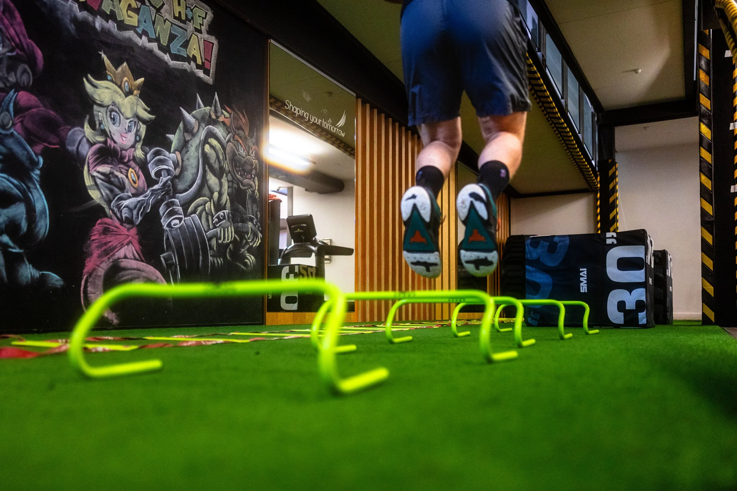 Person exercising at gym with green agility hurdles, colorful wall art featuring cartoon characters, and gym equipment in background.