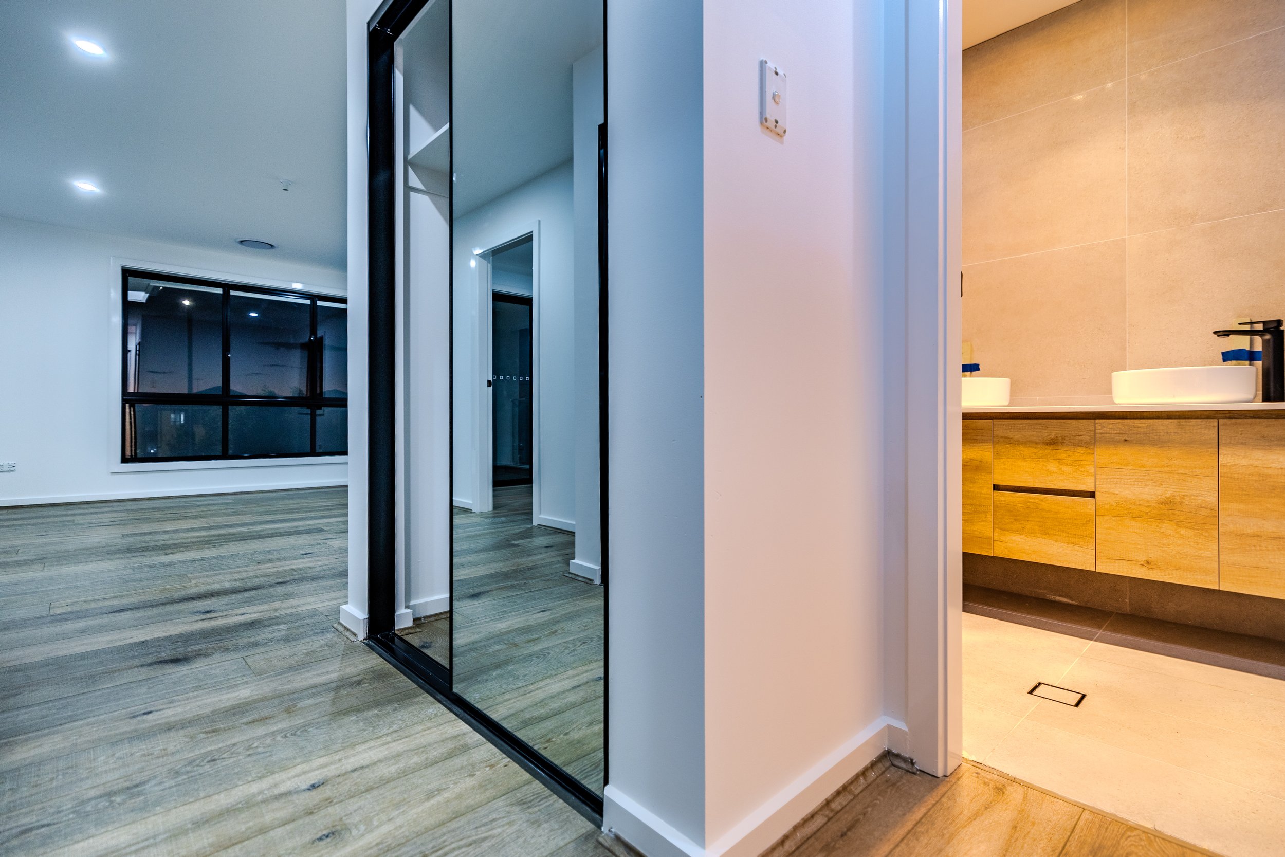 Interior view of a modern apartment with a hardwood floor, large window, and a wall-mounted mirror near a bathroom vanity with dual sinks.