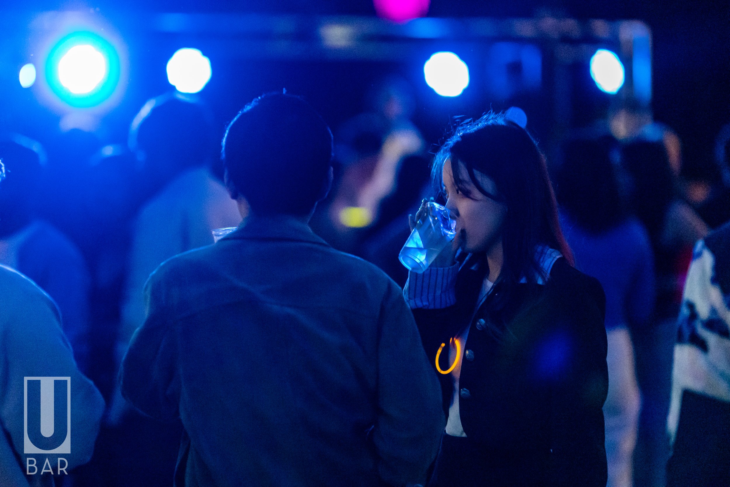 A woman drinking a beverage at a bar or nightclub with blue lighting, talking to a man, with people in the background.