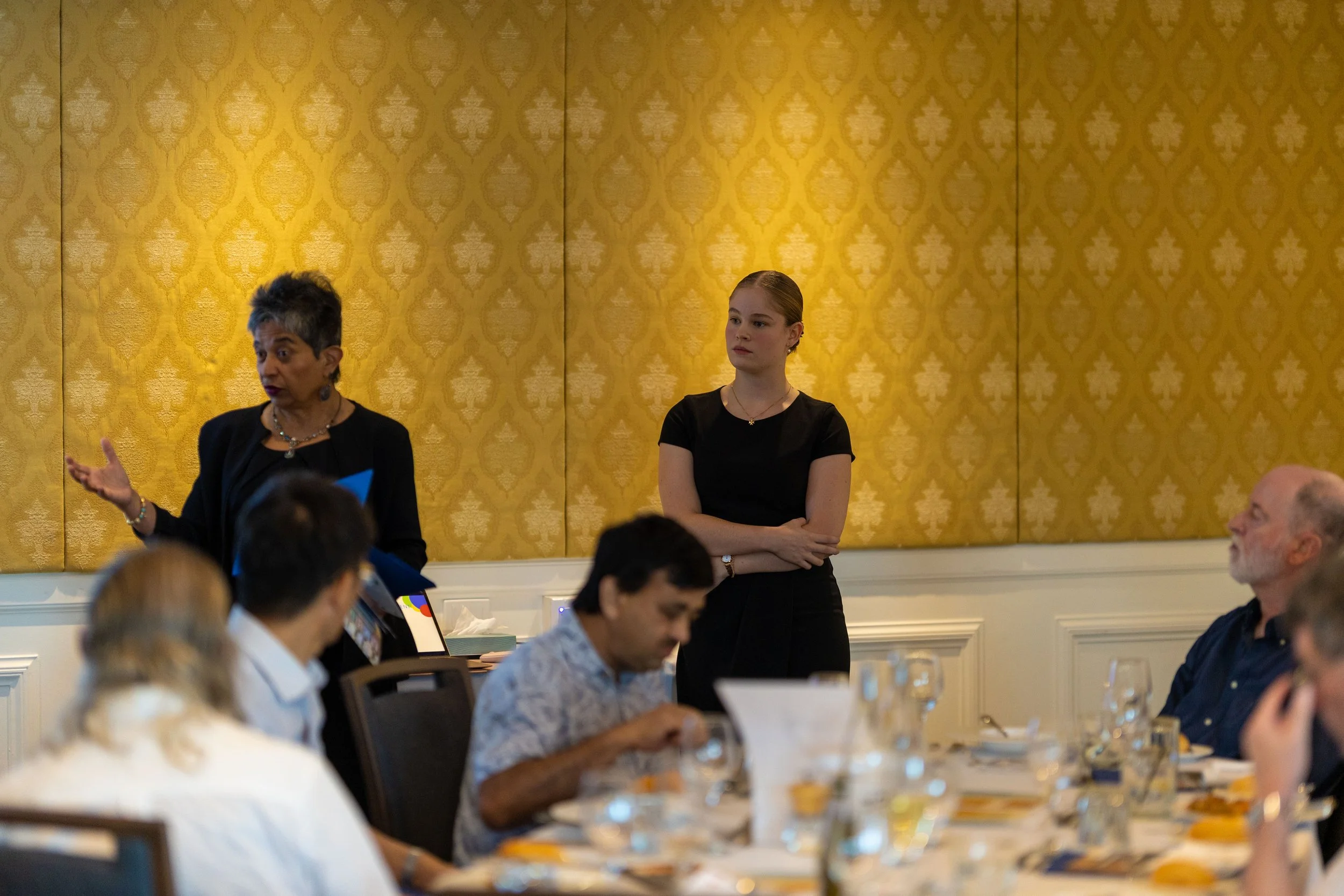 A woman with short gray hair gestures while speaking at a formal event, with a young woman in black standing nearby and other people seated at a dining table.