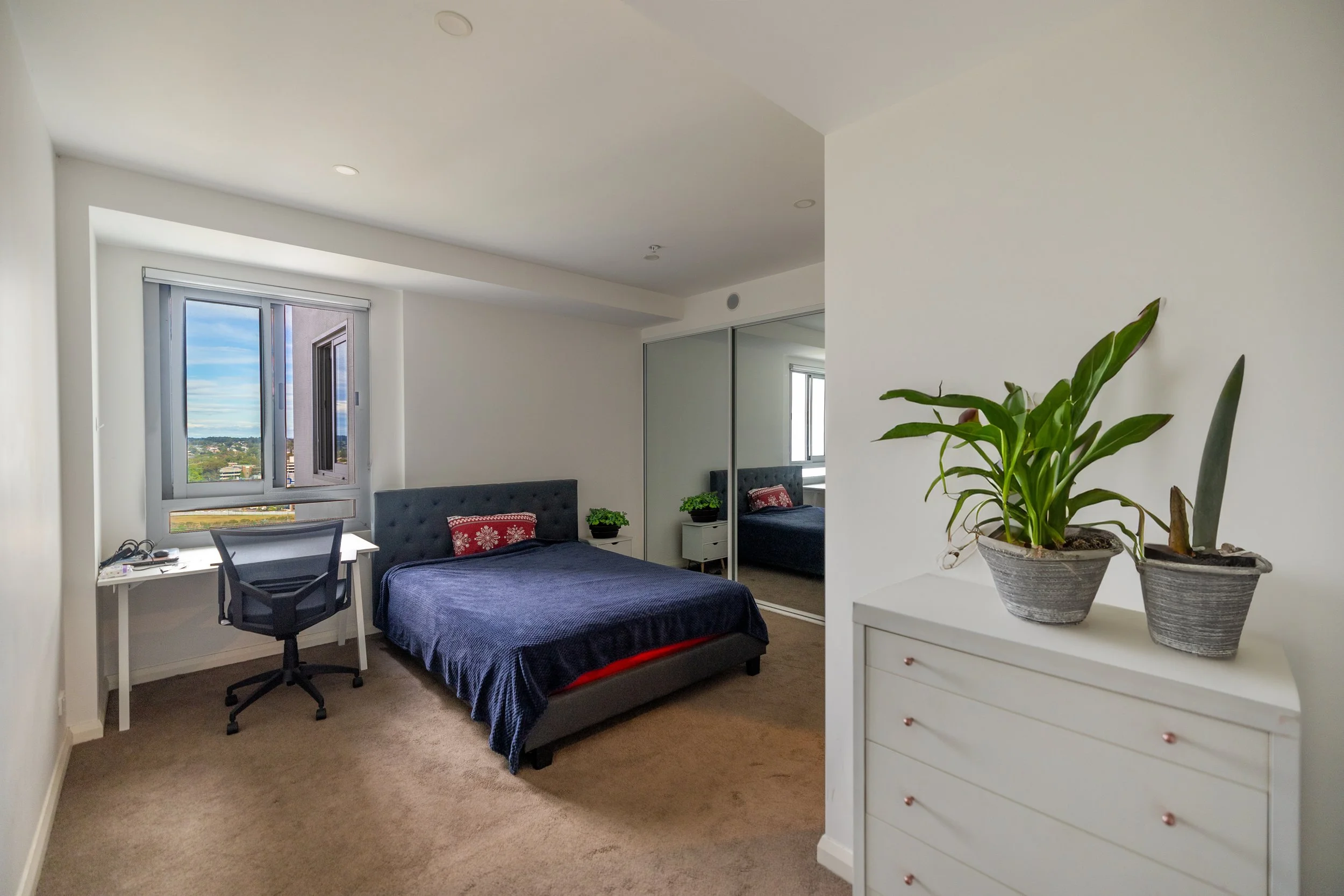 Bedroom with a bed, window, desk, dresser with potted plants, and mirrored closet doors