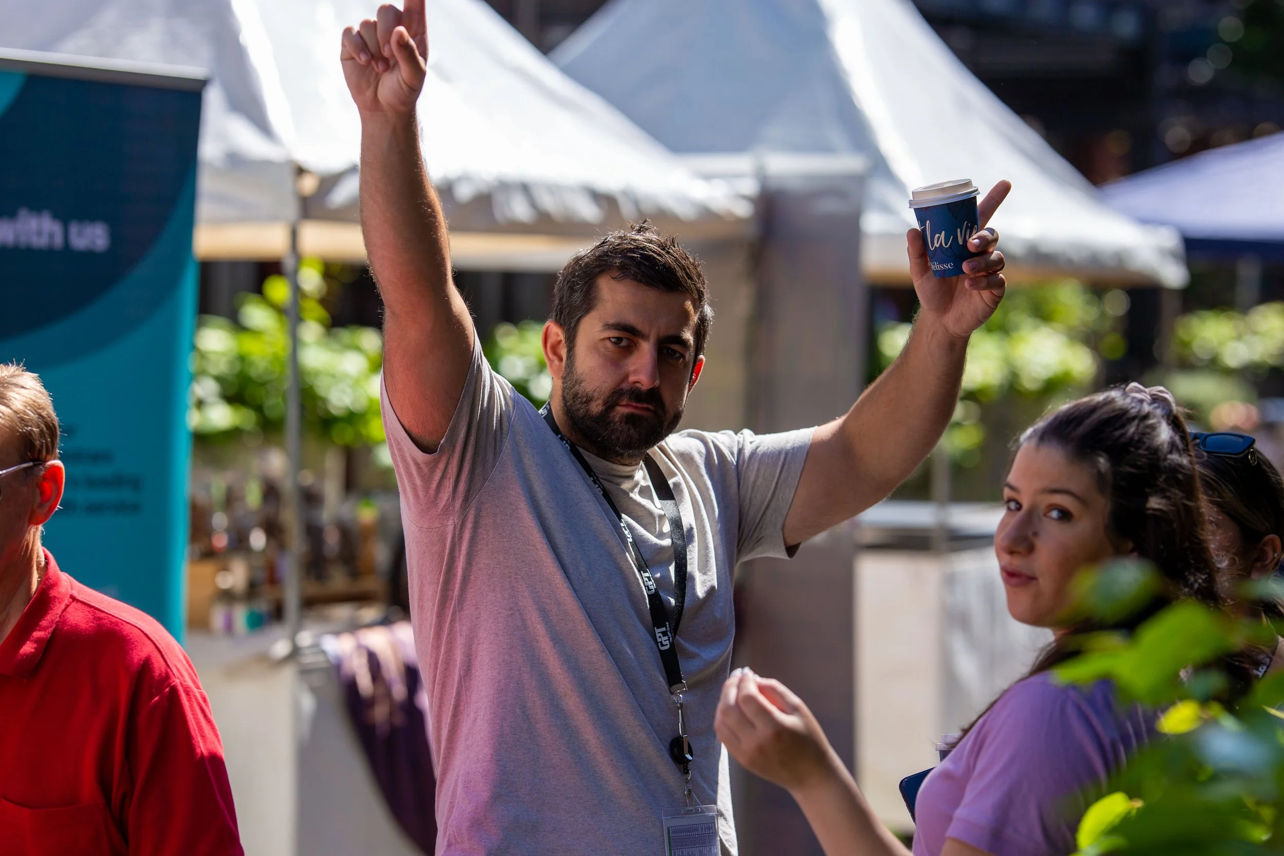 A man with a beard and gray shirt raising his hand at an outdoor event, holding a coffee cup, with two women near him under tents and greenery in the background.