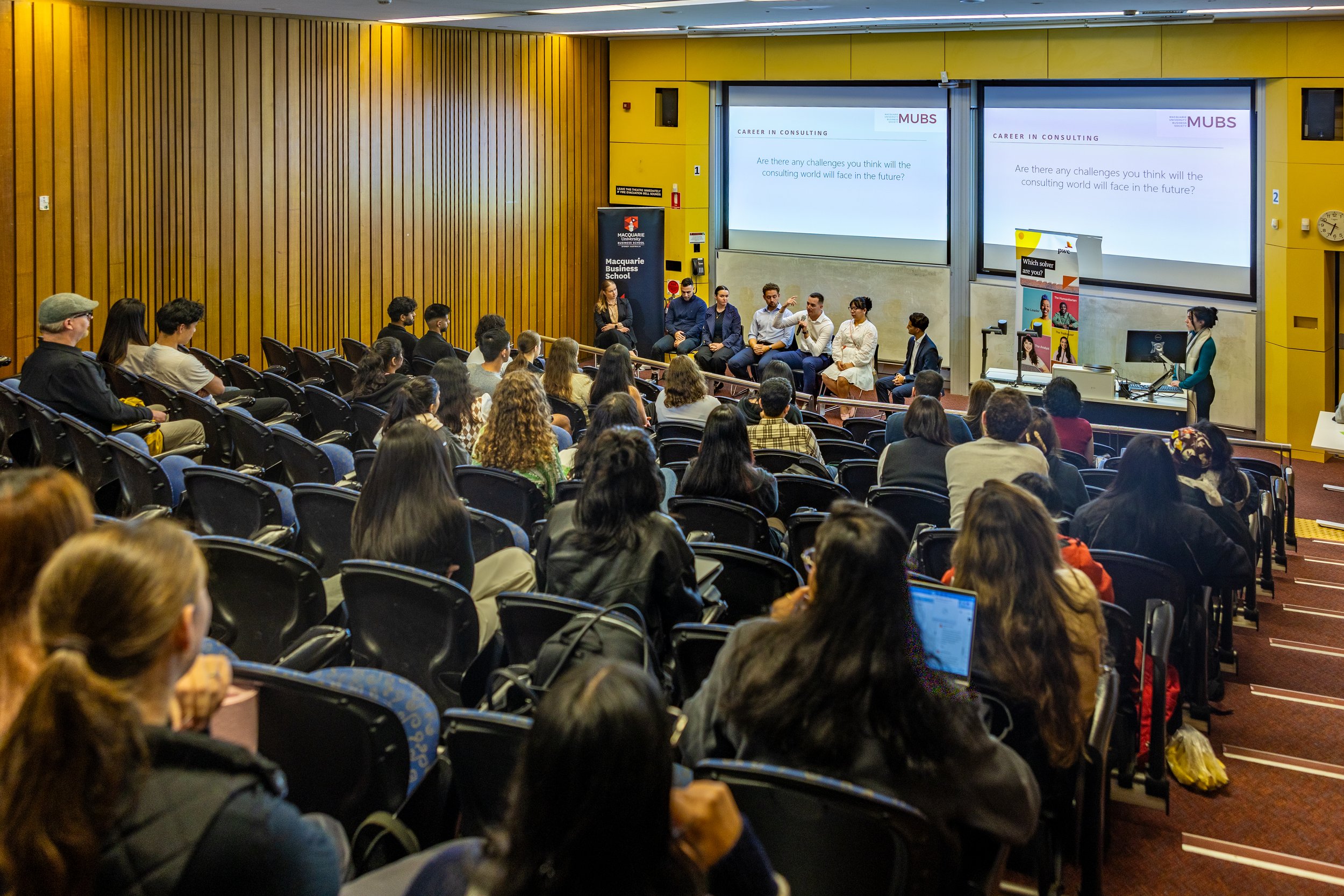 A panel discussion at a university conference with an audience seated in rows, and seven panelists sitting on stage in front of two large screens displaying a presentation slide about challenges in consulting.