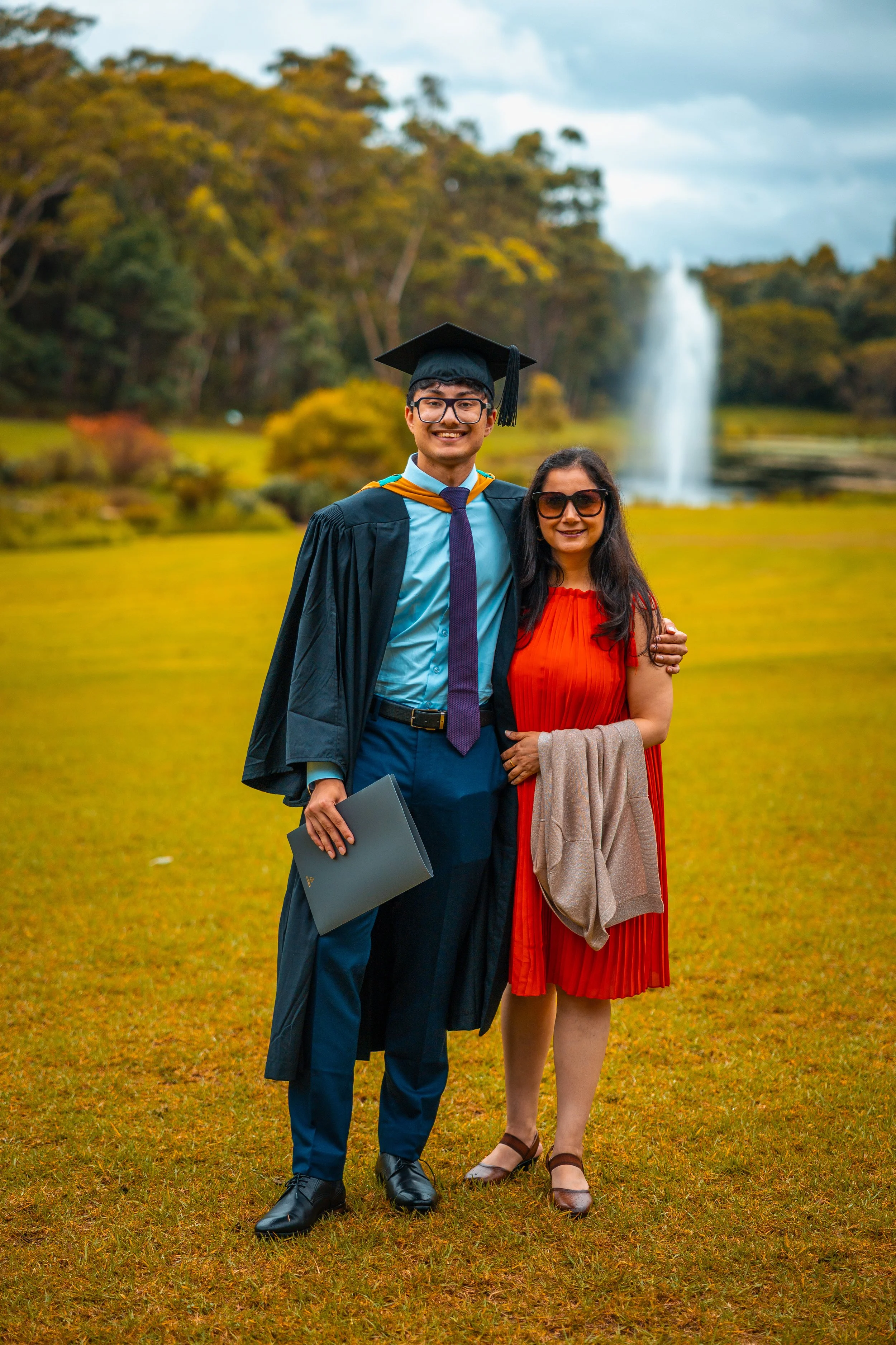 A young man in graduation attire and a woman in a red dress standing outdoors on a grassy area, smiling, with a fountain and trees in the background.