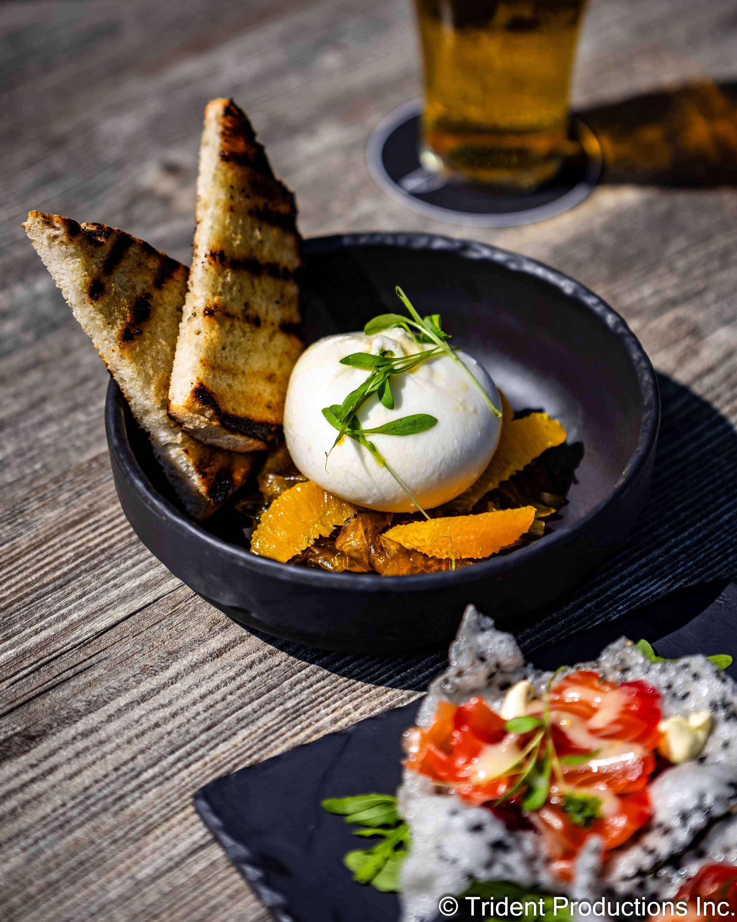 A black bowl with grilled bread, a scoop of burrata cheese garnished with greens, and orange slices, on a rustic wooden table with a glass of beer in the background.