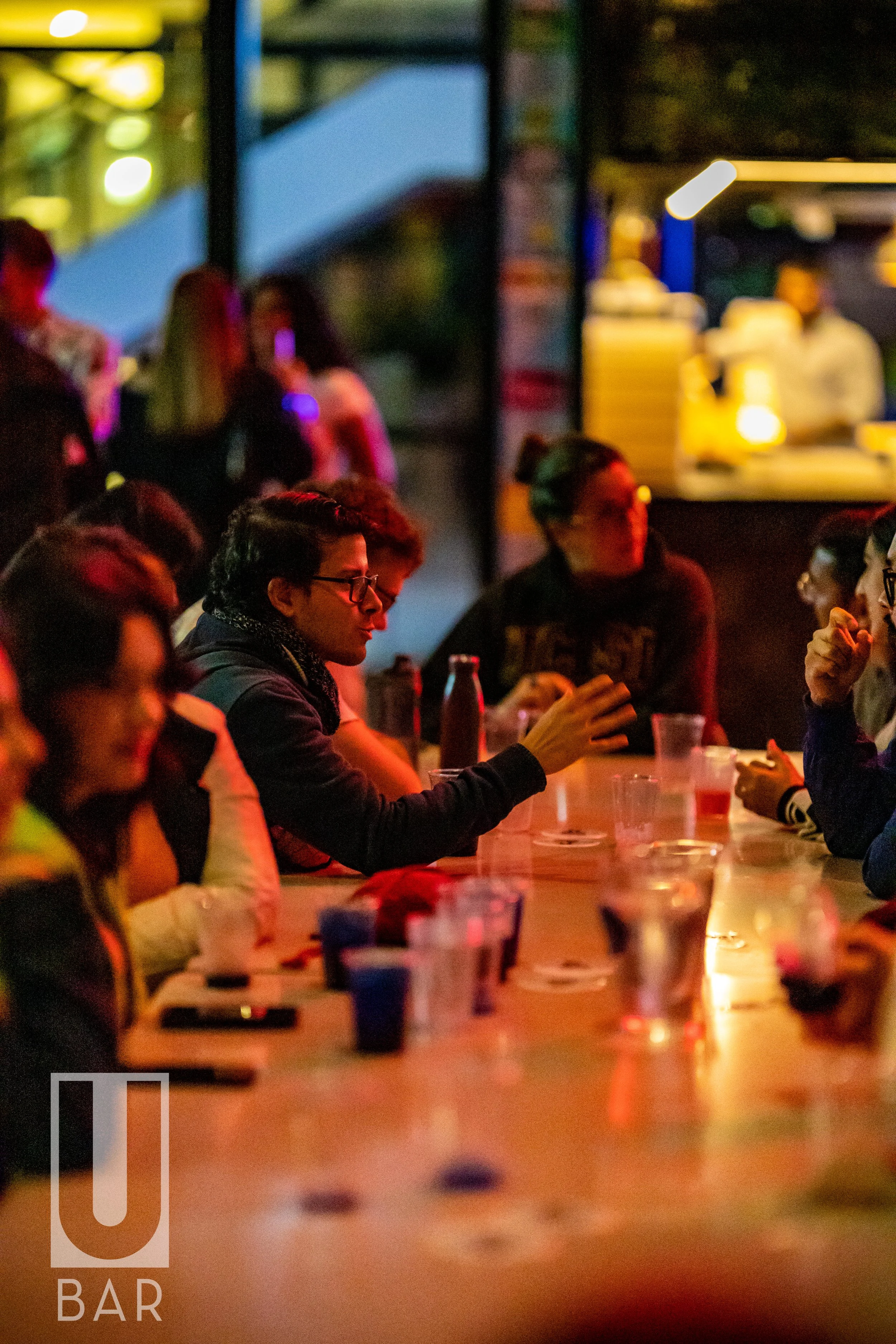 People sitting at a bar table under warm lighting, some drinking, with a bartender in the background and a logo 'U BAR' in the corner.