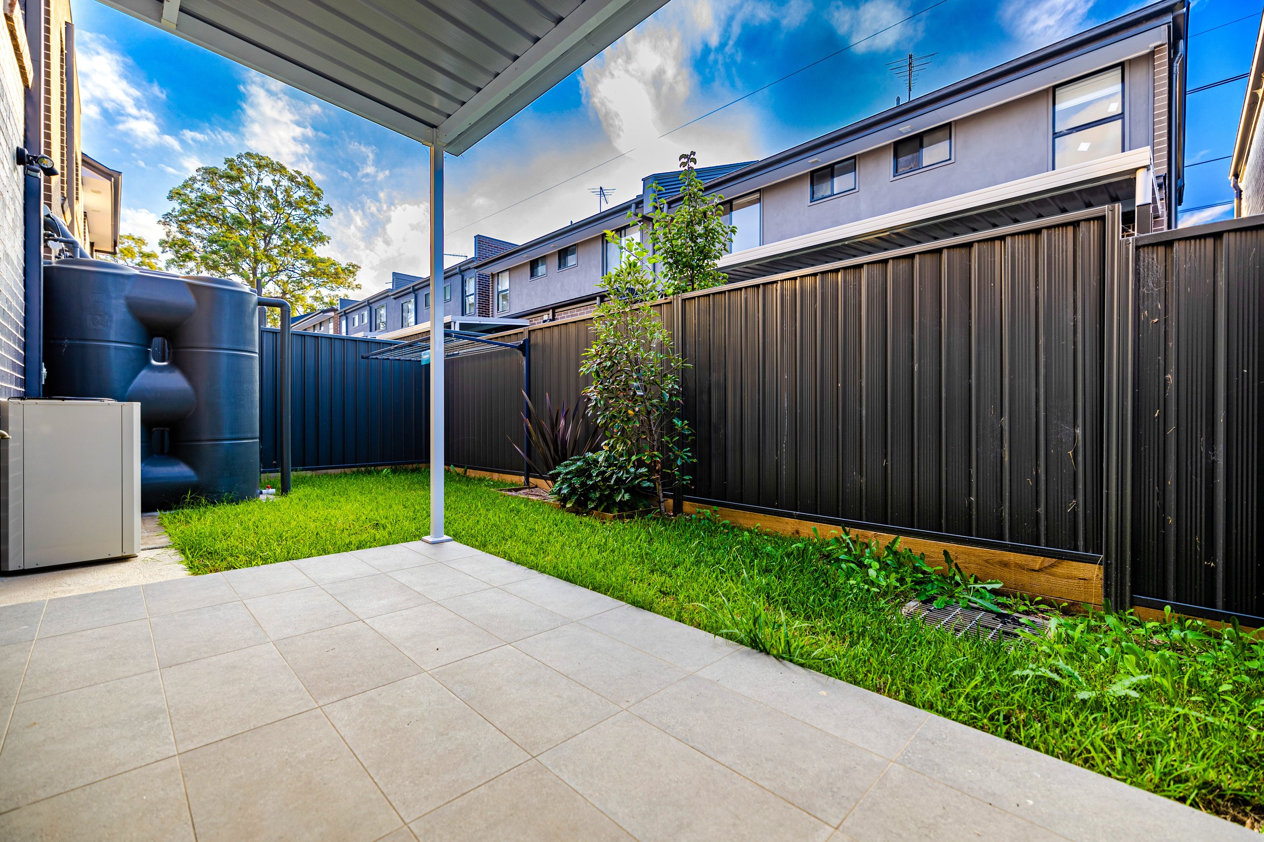 Backyard patio with tiled floor, grass patch, small tree, black metal fence, and a large water tank in the corner. Residential buildings are visible in the background under a partly cloudy sky.