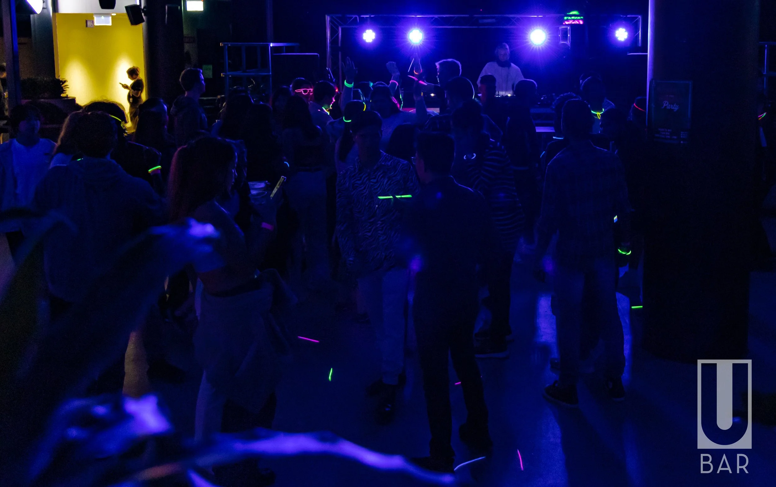 People dancing and socializing at a nightclub or bar with neon and UV lighting, with a DJ or performer on stage in the background.