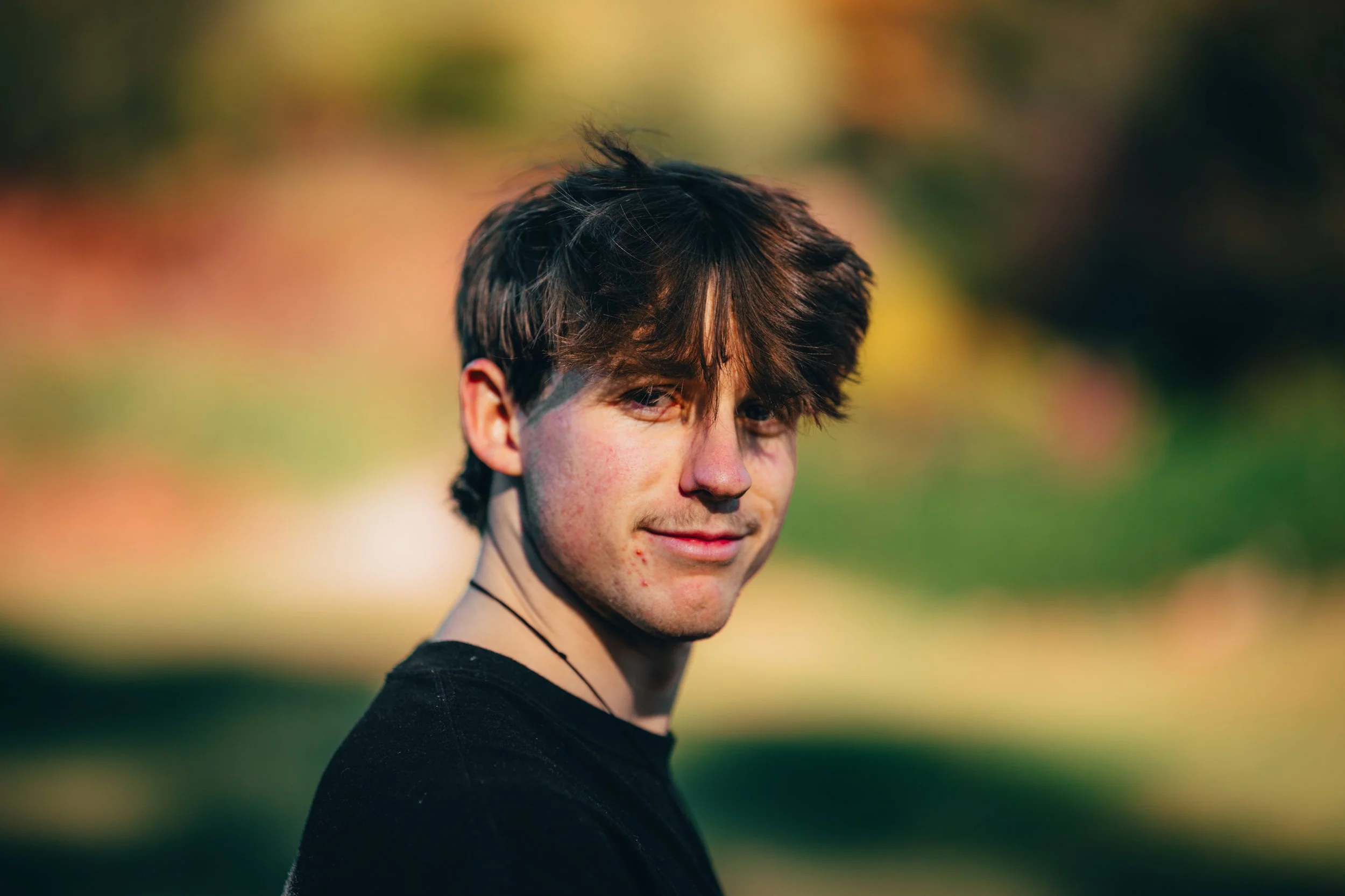 A young man with dark brown hair and light skin outdoors in sunlight, wearing a black shirt and a thin necklace, with a blurred natural background.