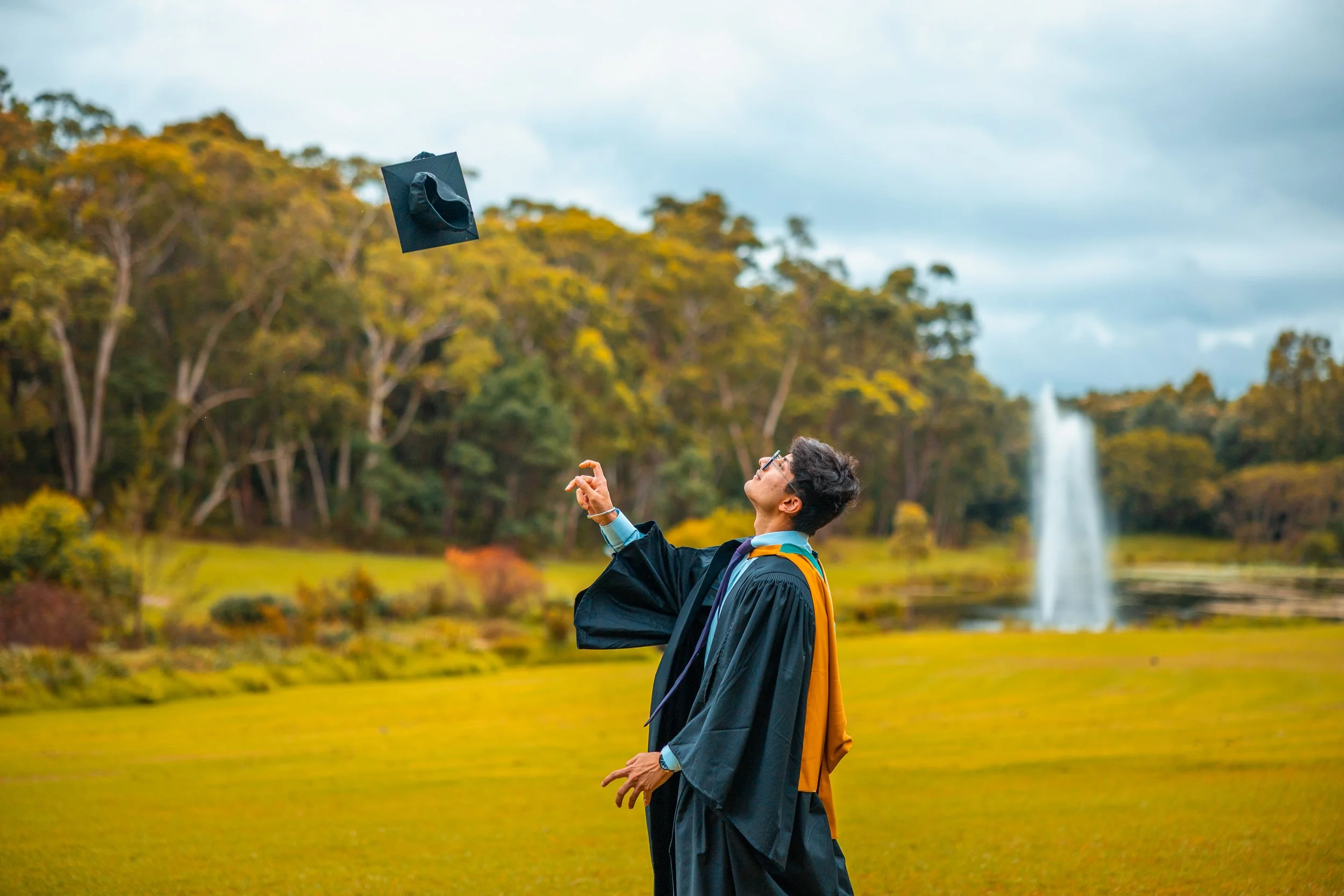 A young man in graduation gown and cap tossing his cap in the air outdoors with a fountain in the background.