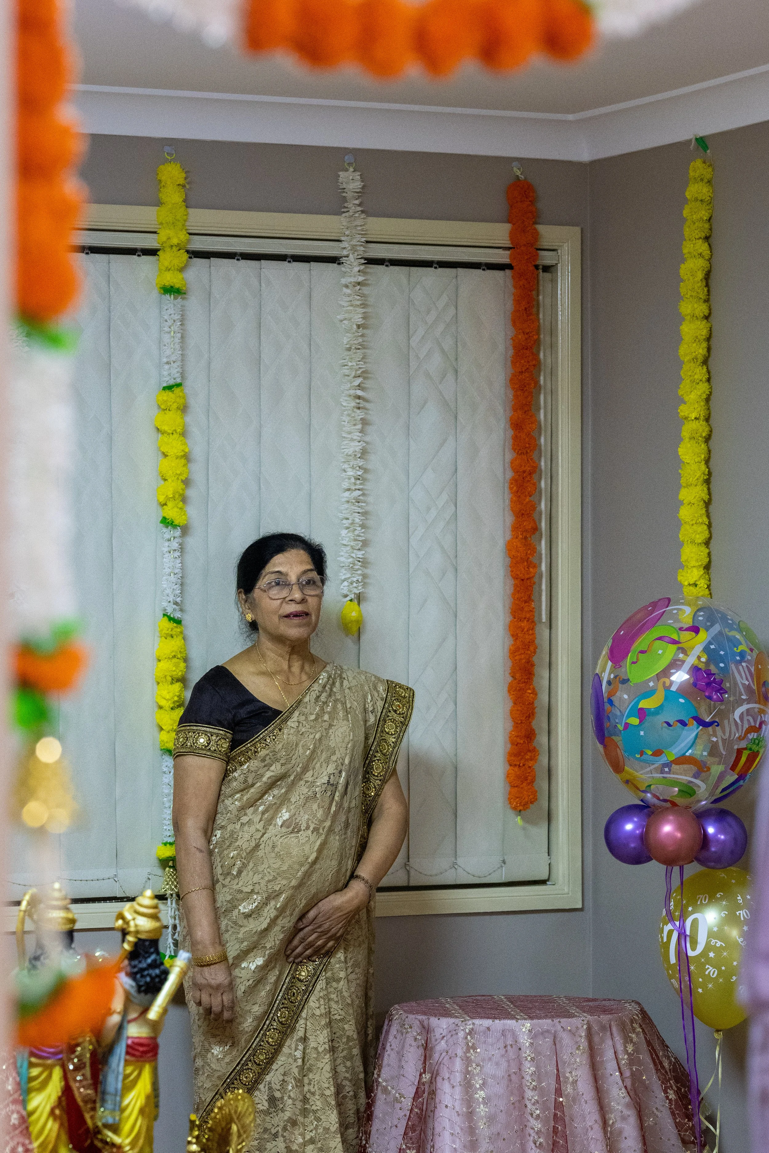 An elderly woman dressed in a gold saree standing in a decorated room for a celebration, with colorful garlands hanging on the wall and balloons on the right side.