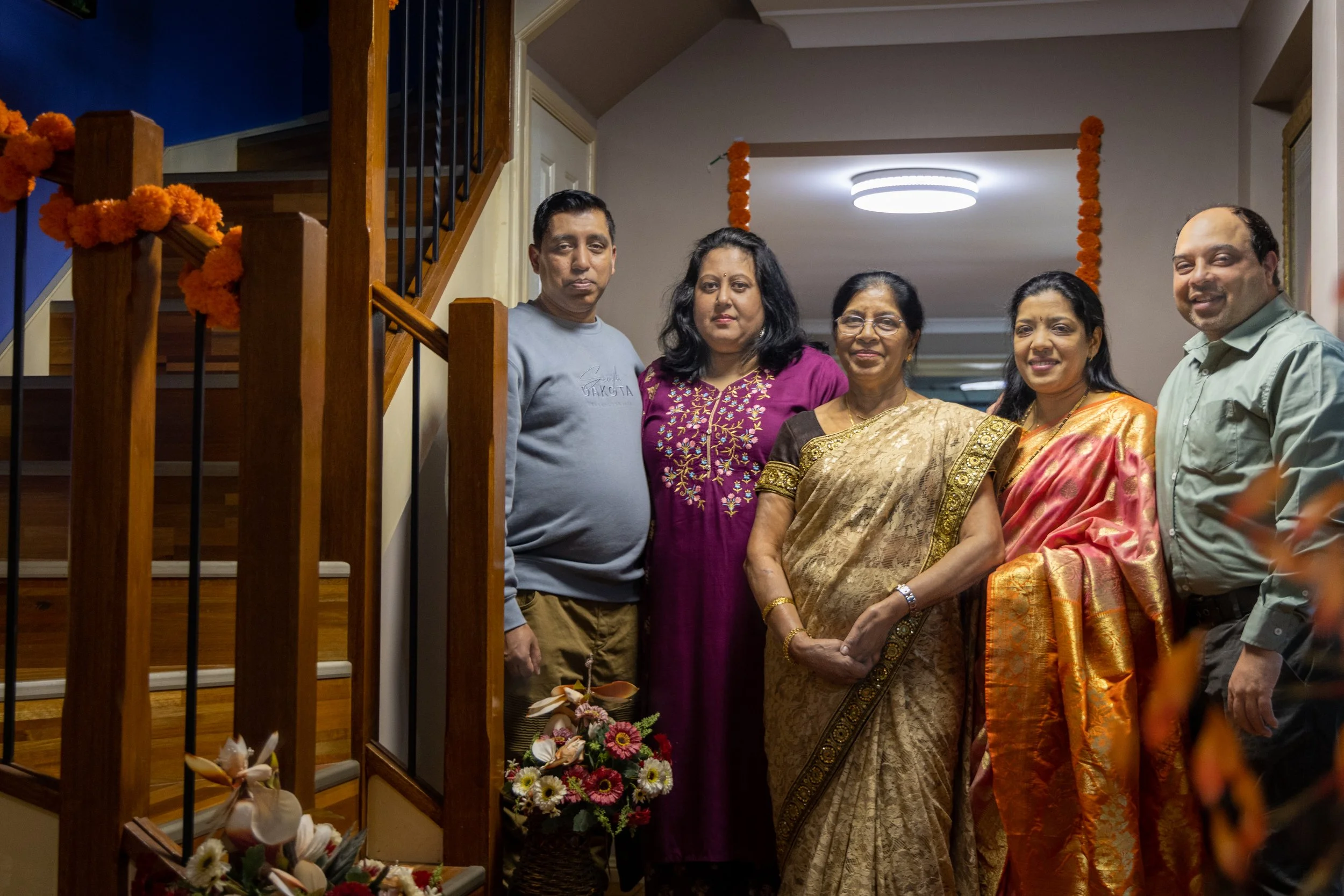Group of six people standing inside a house, dressed in traditional Indian clothing, posing for a photo with floral decorations in the background.