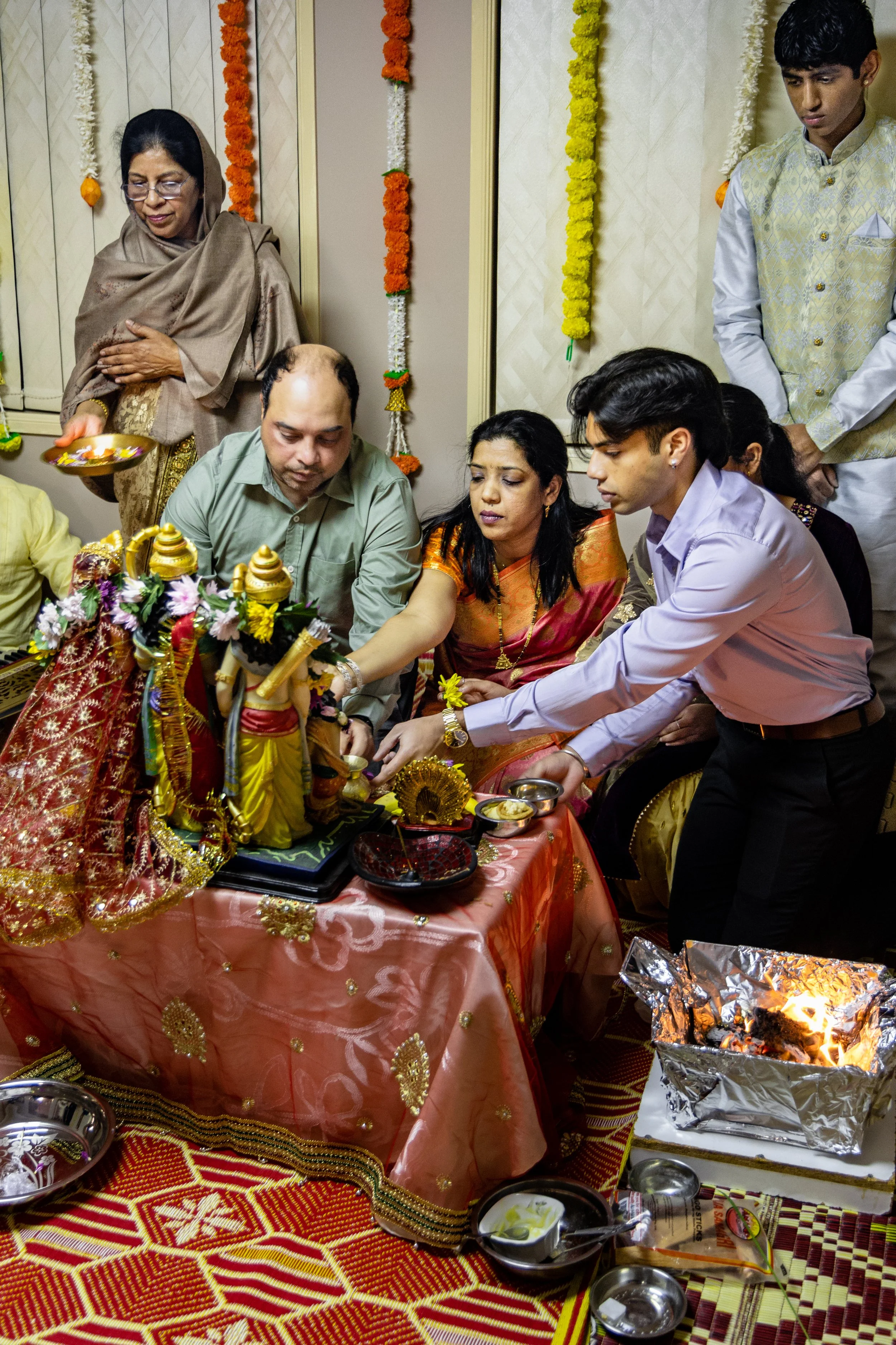 Indian family participating in a religious ritual with idols of Lord Krishna and Radha, offering prayers, in a decorated room during a festive occasion.