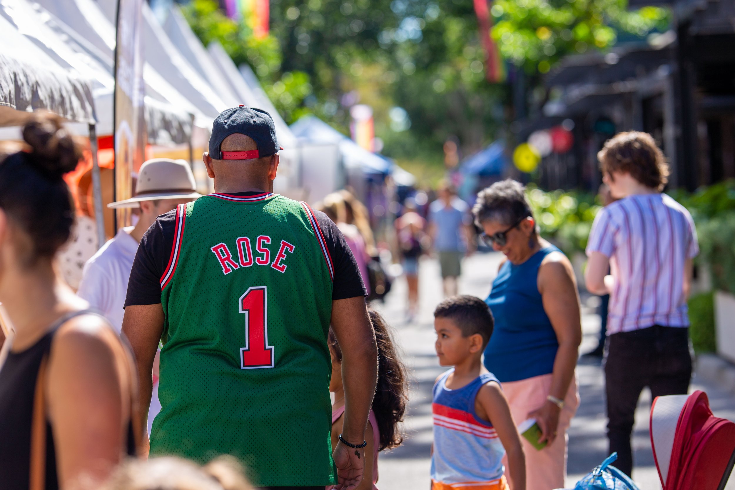 A busy outdoor market scene with people of different ages and styles, some shopping and others walking, on a sunny day with green trees and vendor stalls in the background.