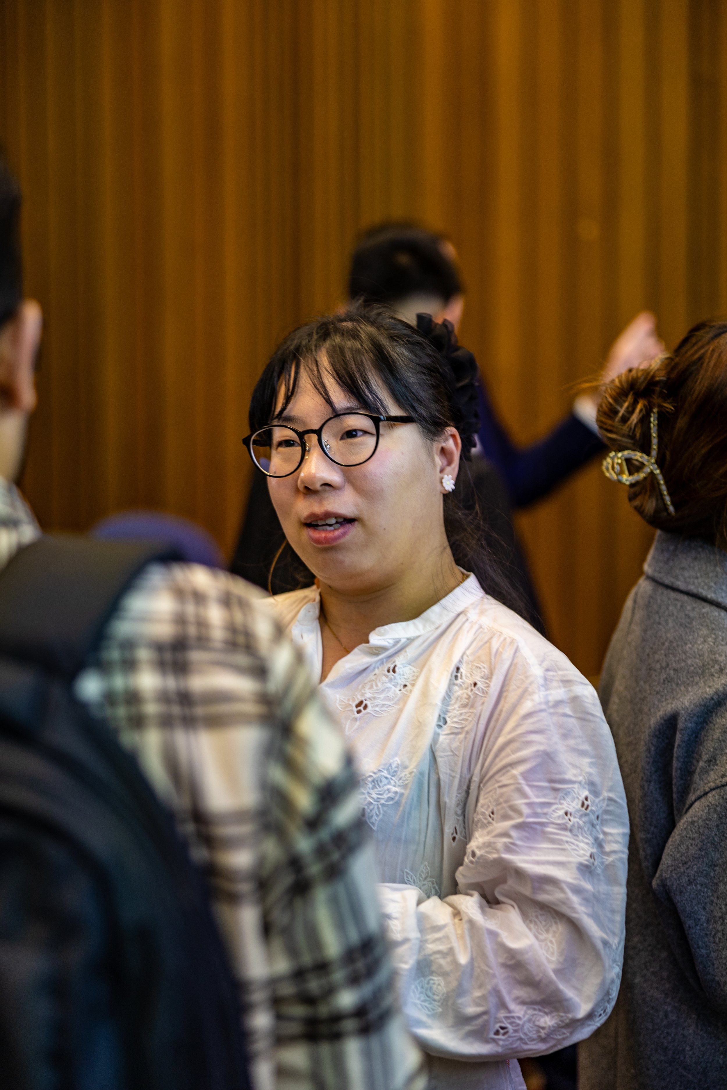 A woman with glasses and earrings engaged in conversation at a social gathering, with other people around her and a wooden wall in the background.