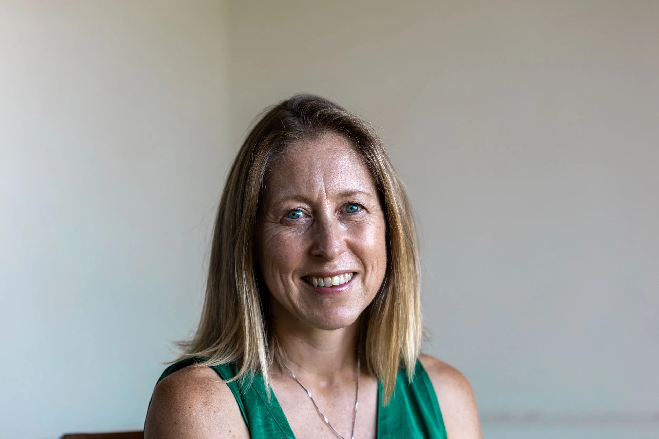 A smiling woman with shoulder-length blonde hair, wearing a green sleeveless top, sitting indoors against a plain light-colored wall.