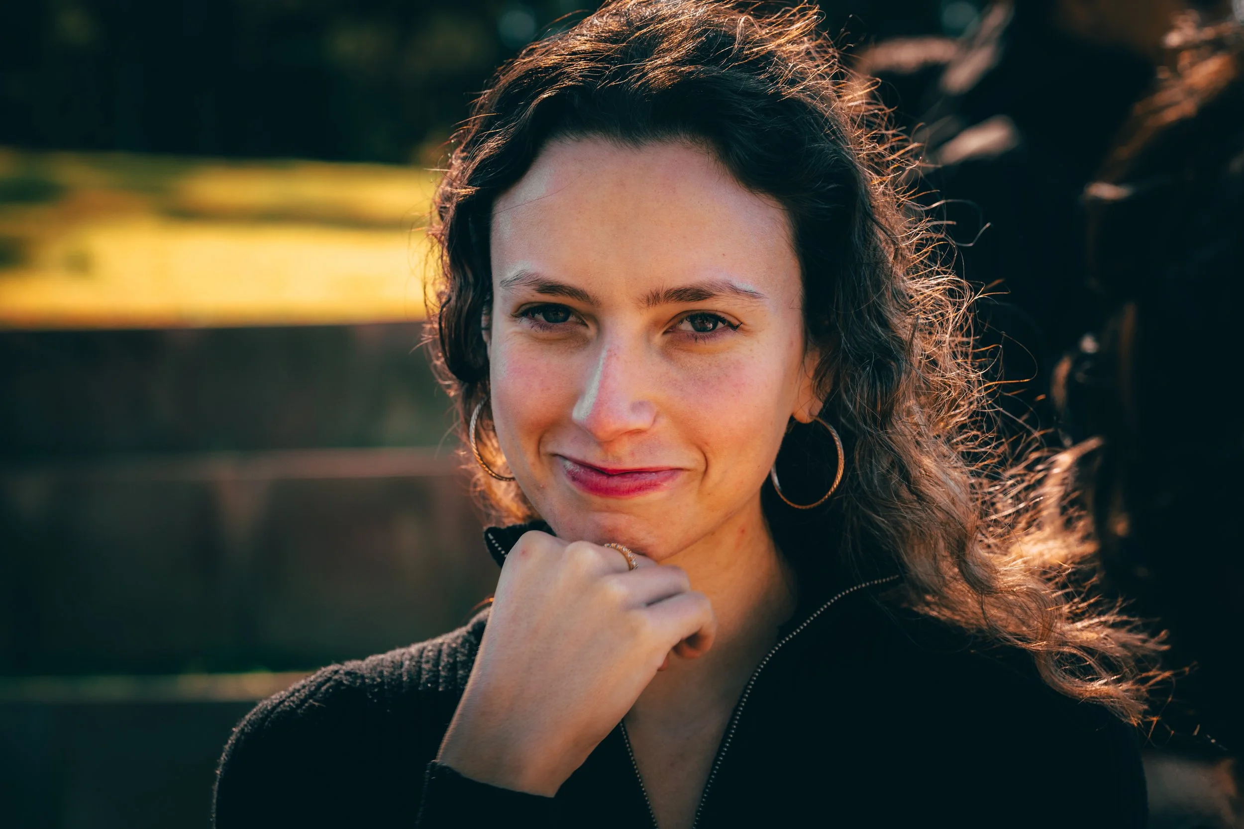 A woman with curly brown hair, wearing hoop earrings and a black zip-up top, smiling outdoors during sunset with natural light illuminating her face.