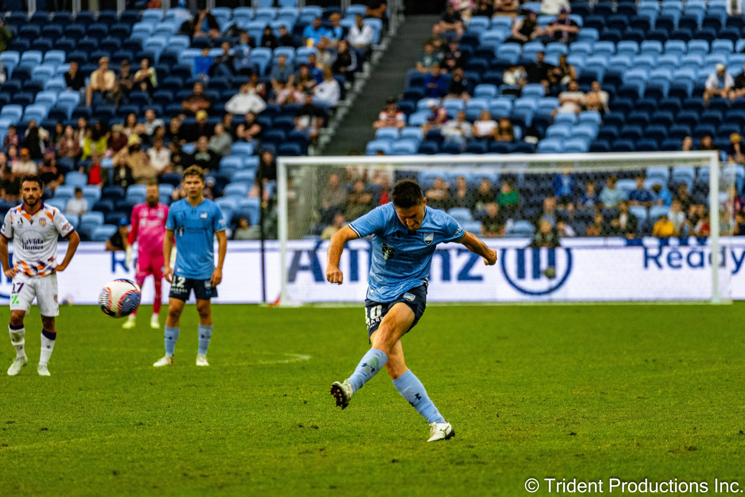 A soccer player in a light blue uniform kicking a ball on a soccer field during a match, with other players and spectators in the background.