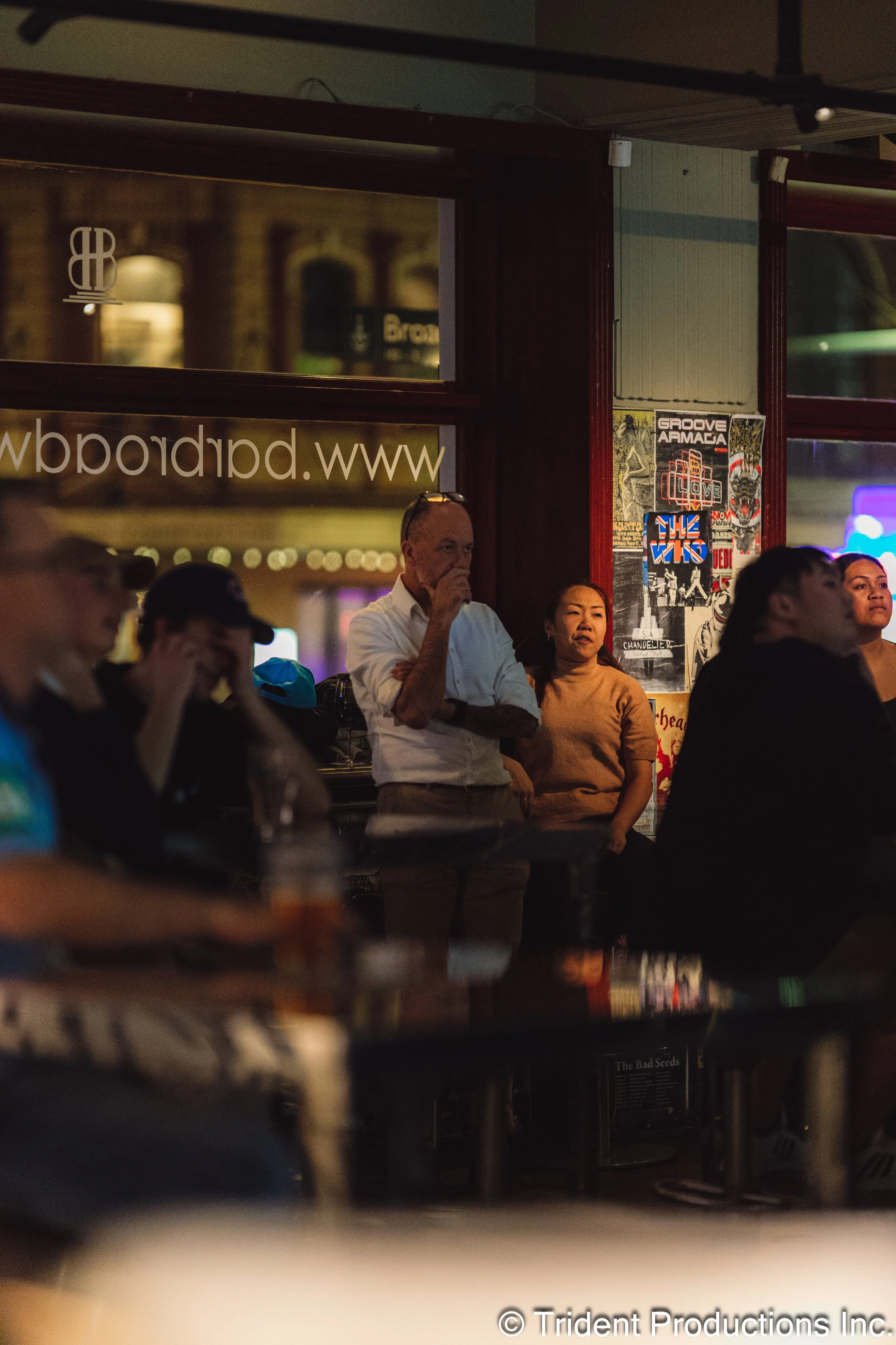People gathered indoors at a dimly lit venue, some seated at tables and others standing near a wall with posters, engaged in watching an event or performance.