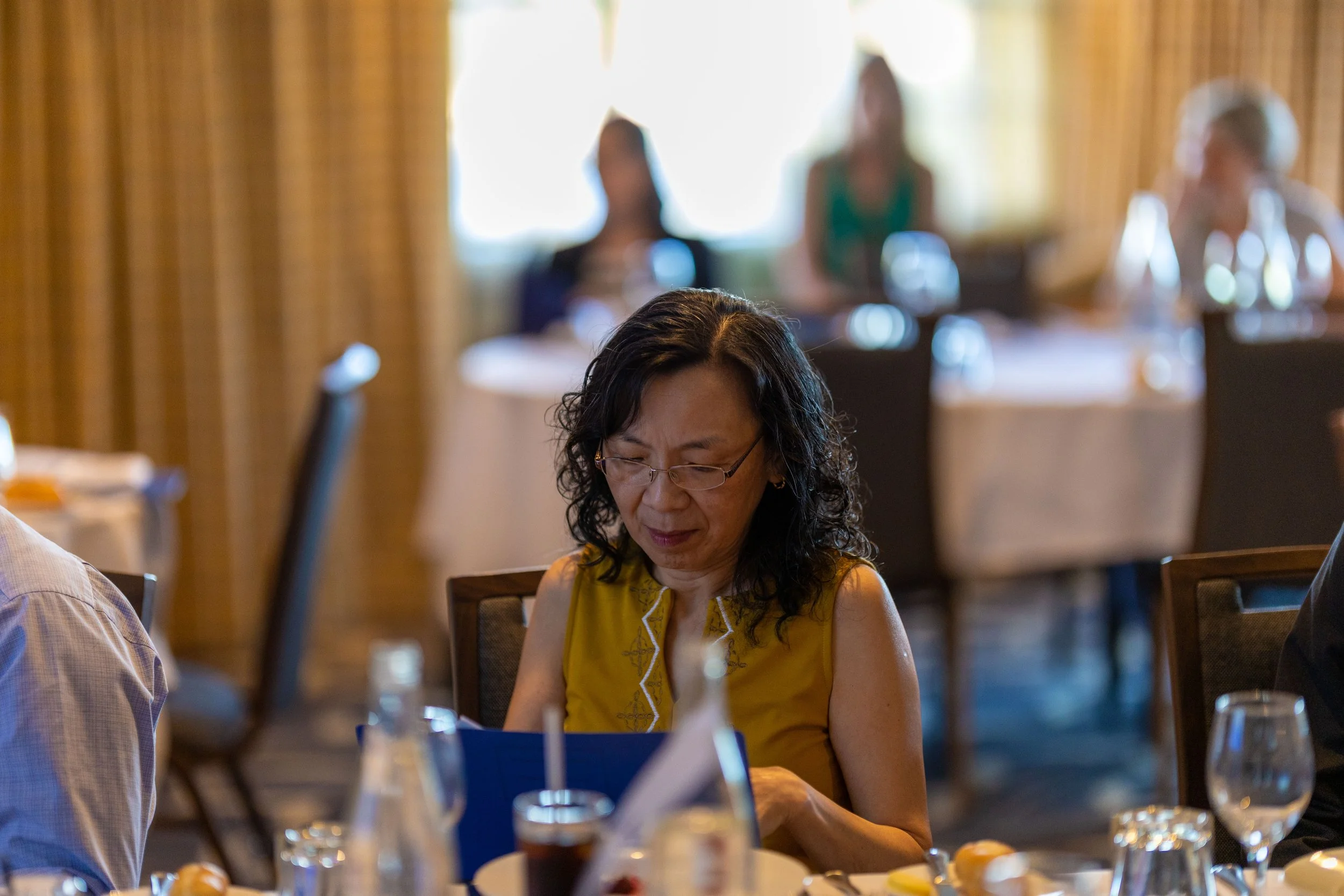 A woman in a yellow top sitting at a dining table, looking at a menu, in a restaurant with other diners in the background.