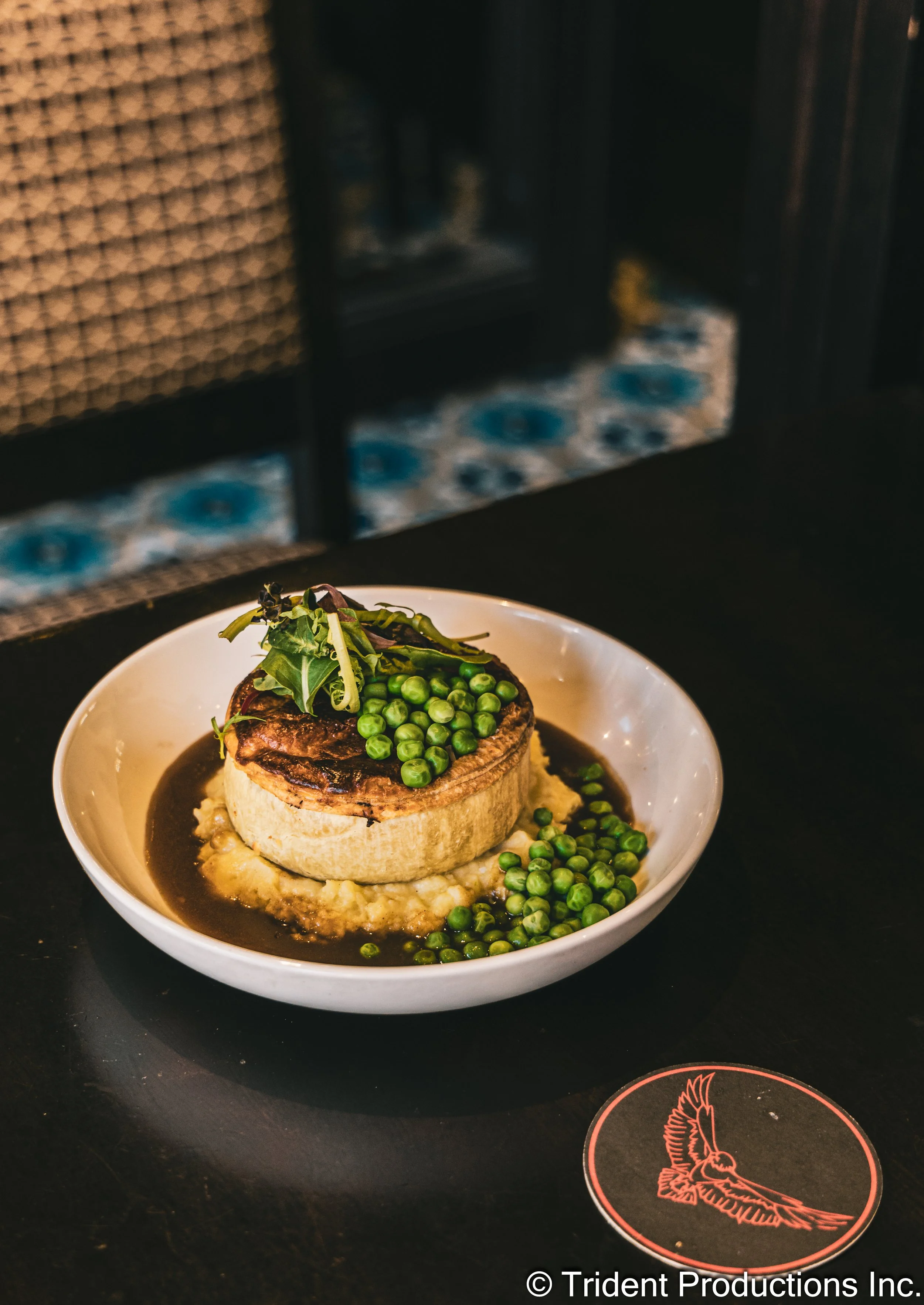 A plate with a cooked meat patty topped with green peas and salad greens, served over mashed potatoes and dark sauce, on a black table with a coaster featuring a bird illustration.