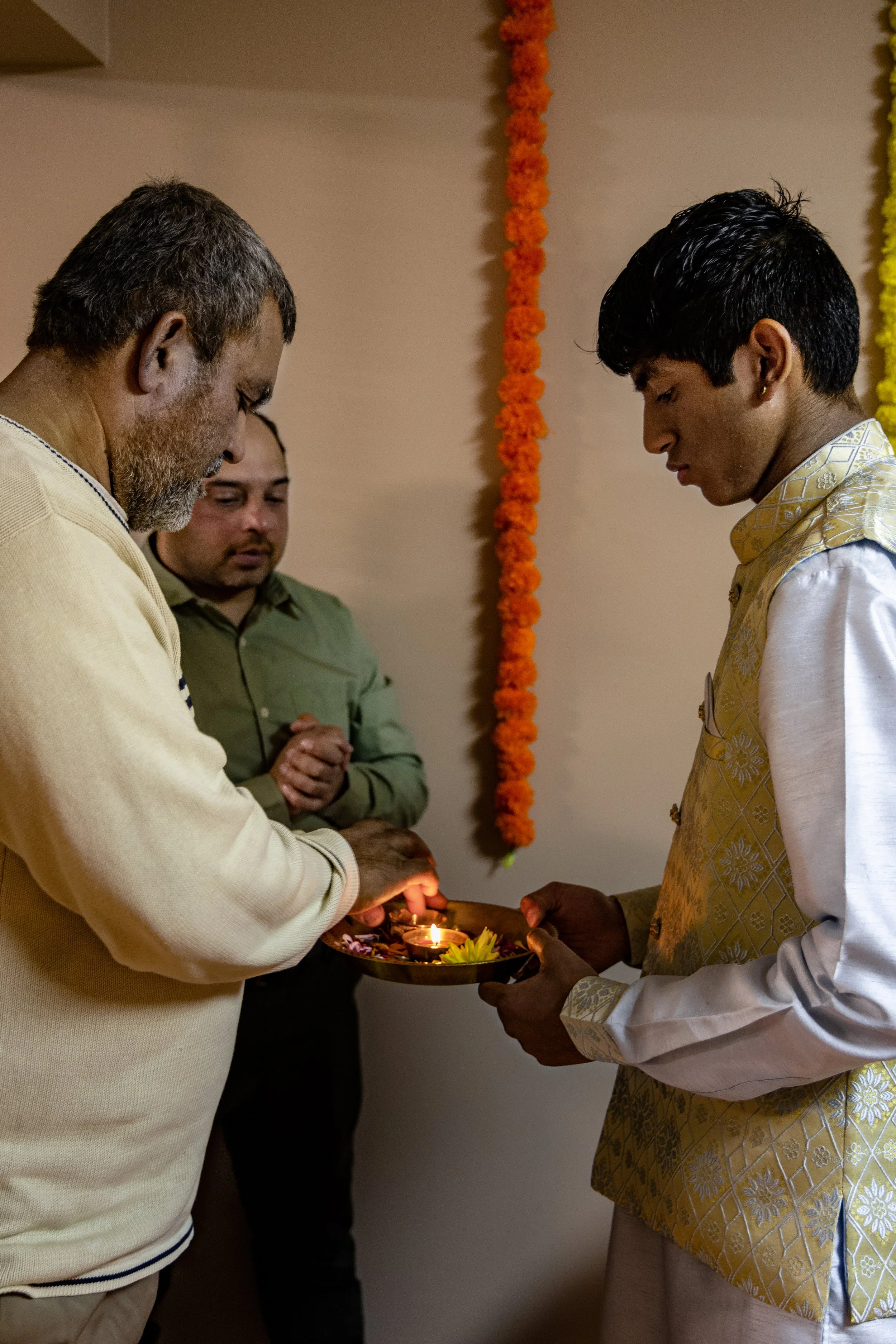 Three men standing indoors during an Indian traditional ceremony, with one lighting a lamp on a plate held by another, while the third looks on, decorated with orange marigold flower garlands on the wall.
